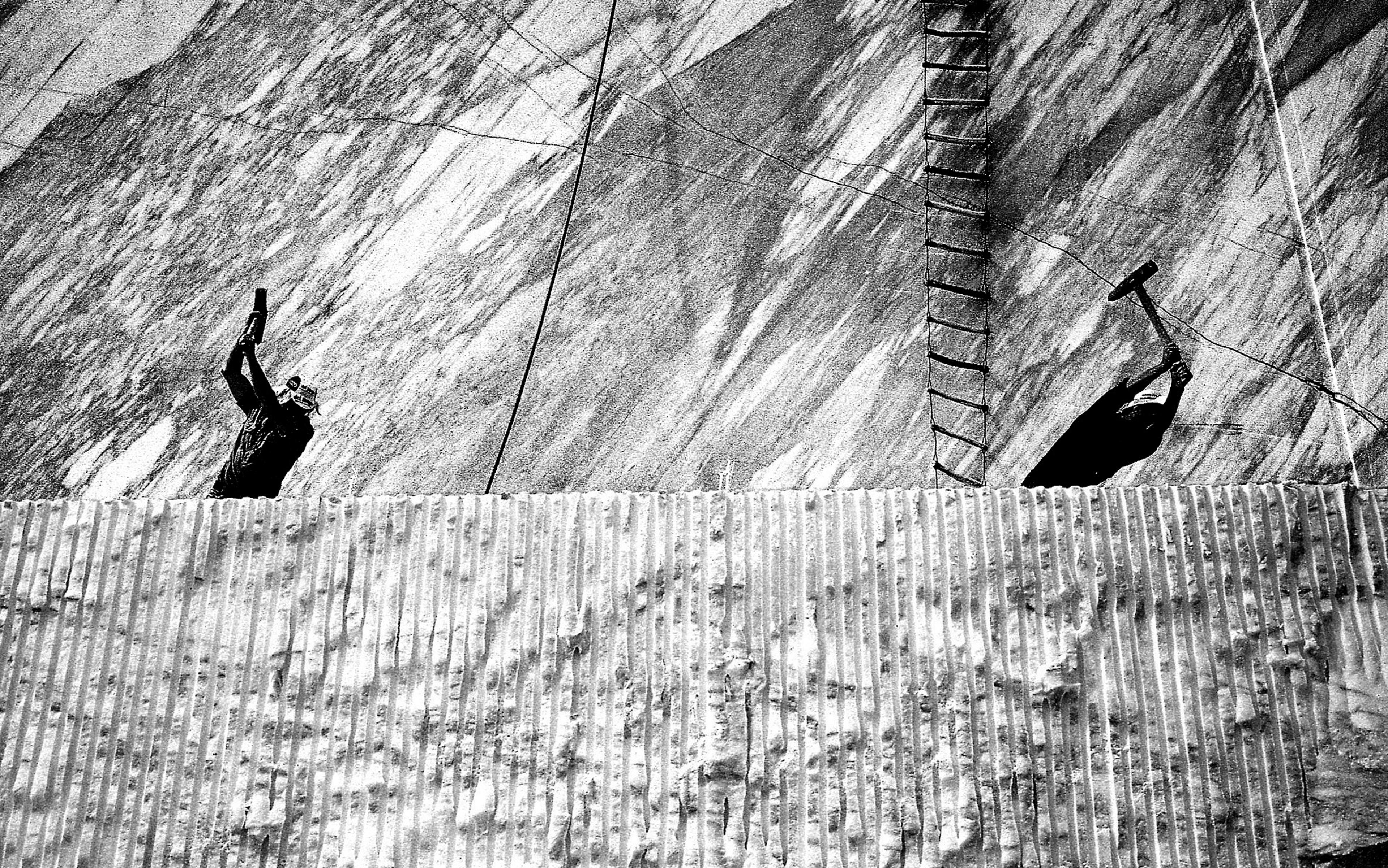 Black and white photo of two silhouetted workers with tools on a rock face. A ladder and cables are seen in the background.