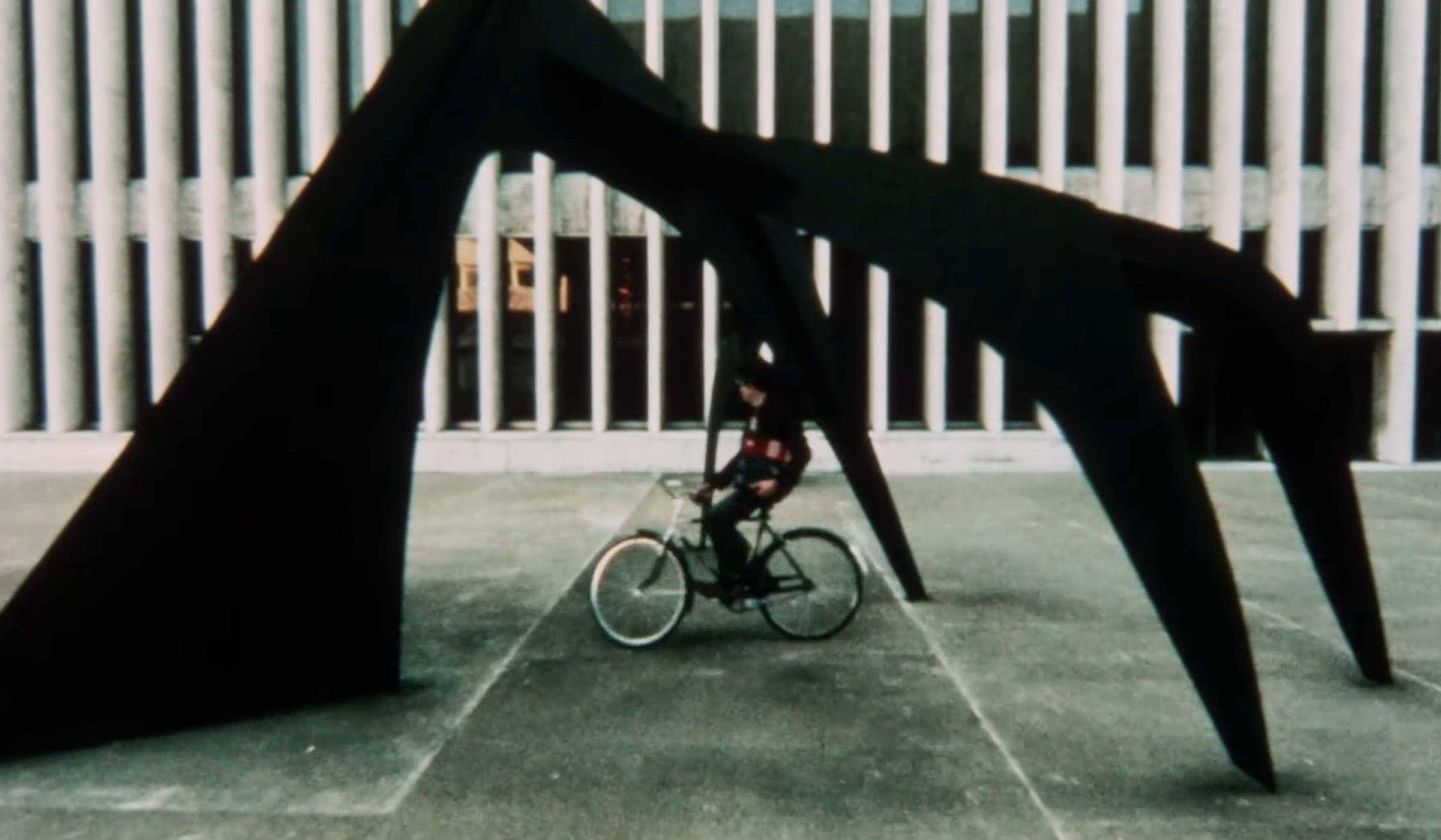 A person on a bicycle under a large, abstract black sculpture in an outdoor plaza with a building featuring vertical columns.