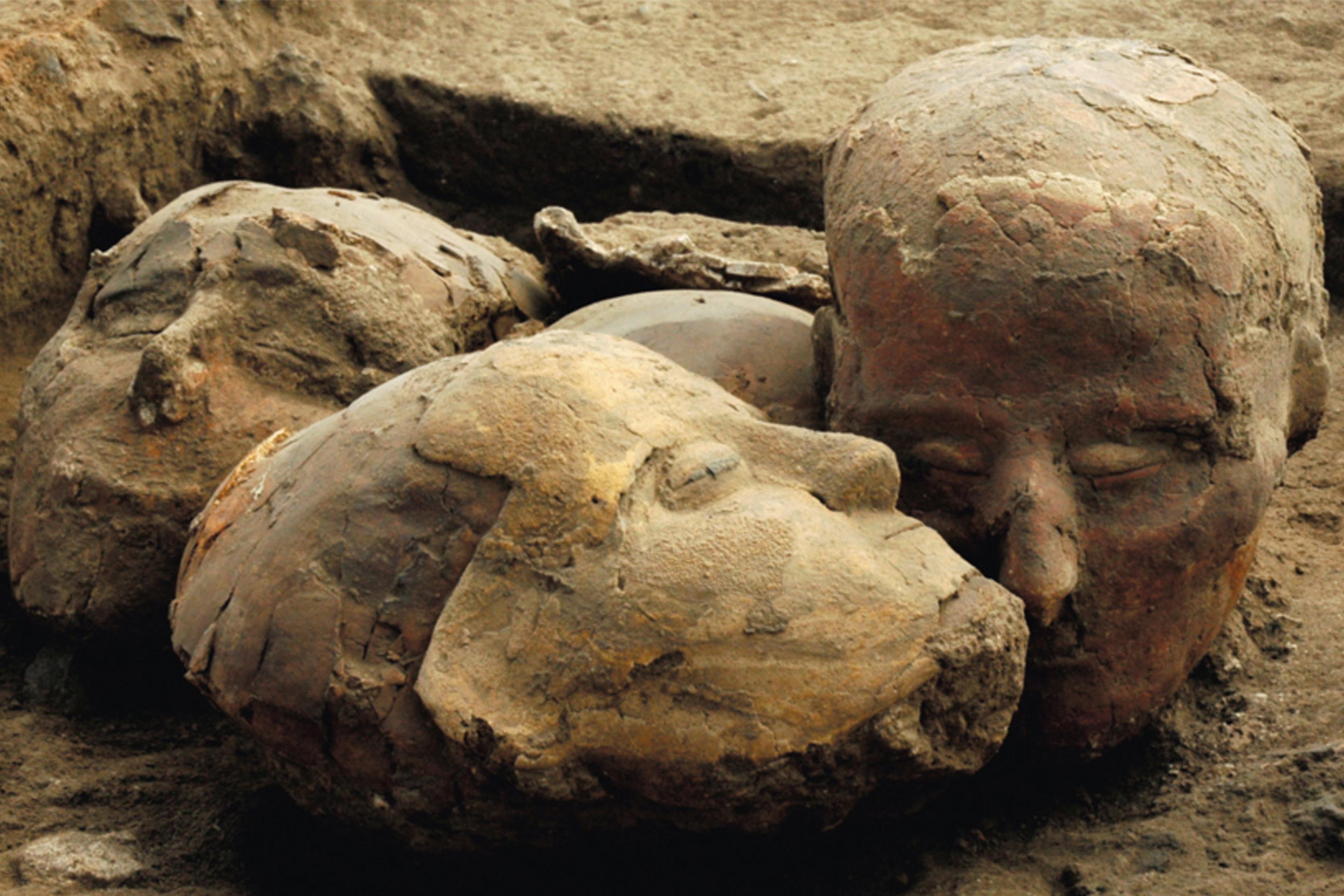 Photo of ancient clay sculptures of human heads partially buried in dirt, showing worn and textured surfaces.