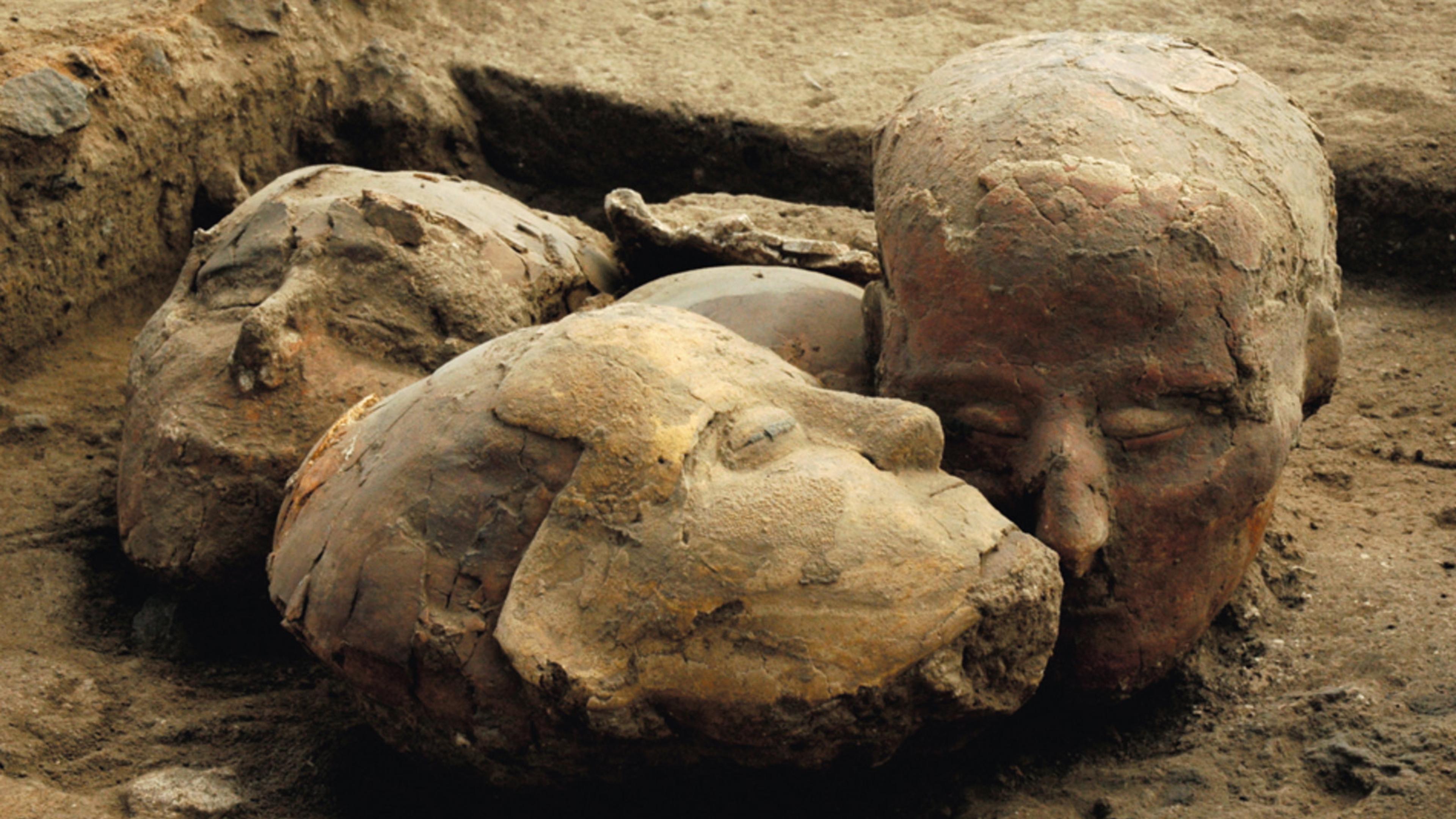 Photo of ancient clay sculptures of human heads partially buried in dirt, showing worn and textured surfaces.