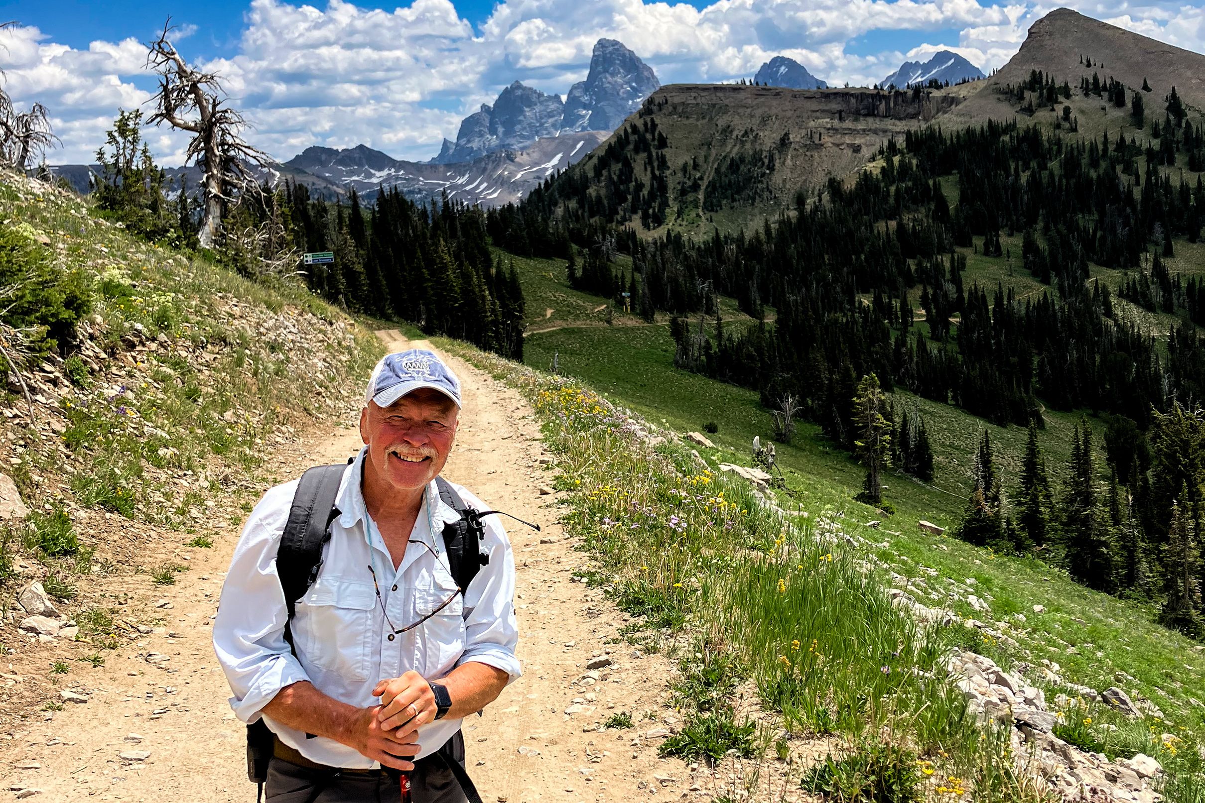 A smiling man hiking on a mountain trail with distant peaks, forests and a bright sky in the background.