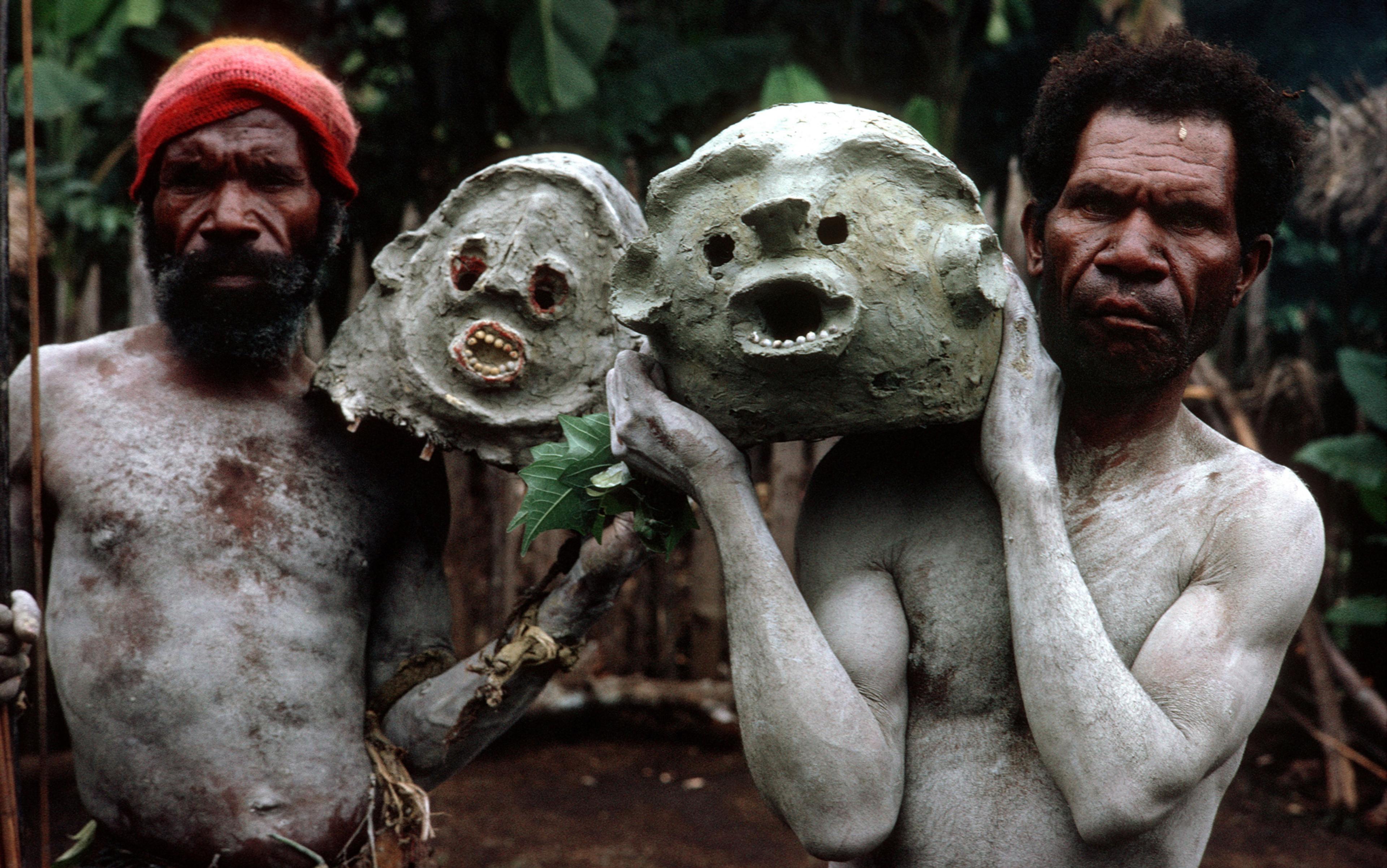 Two Indigenous men in body paint holding clay masks. One wears a red hat. Background shows greenery and a wooden structure.