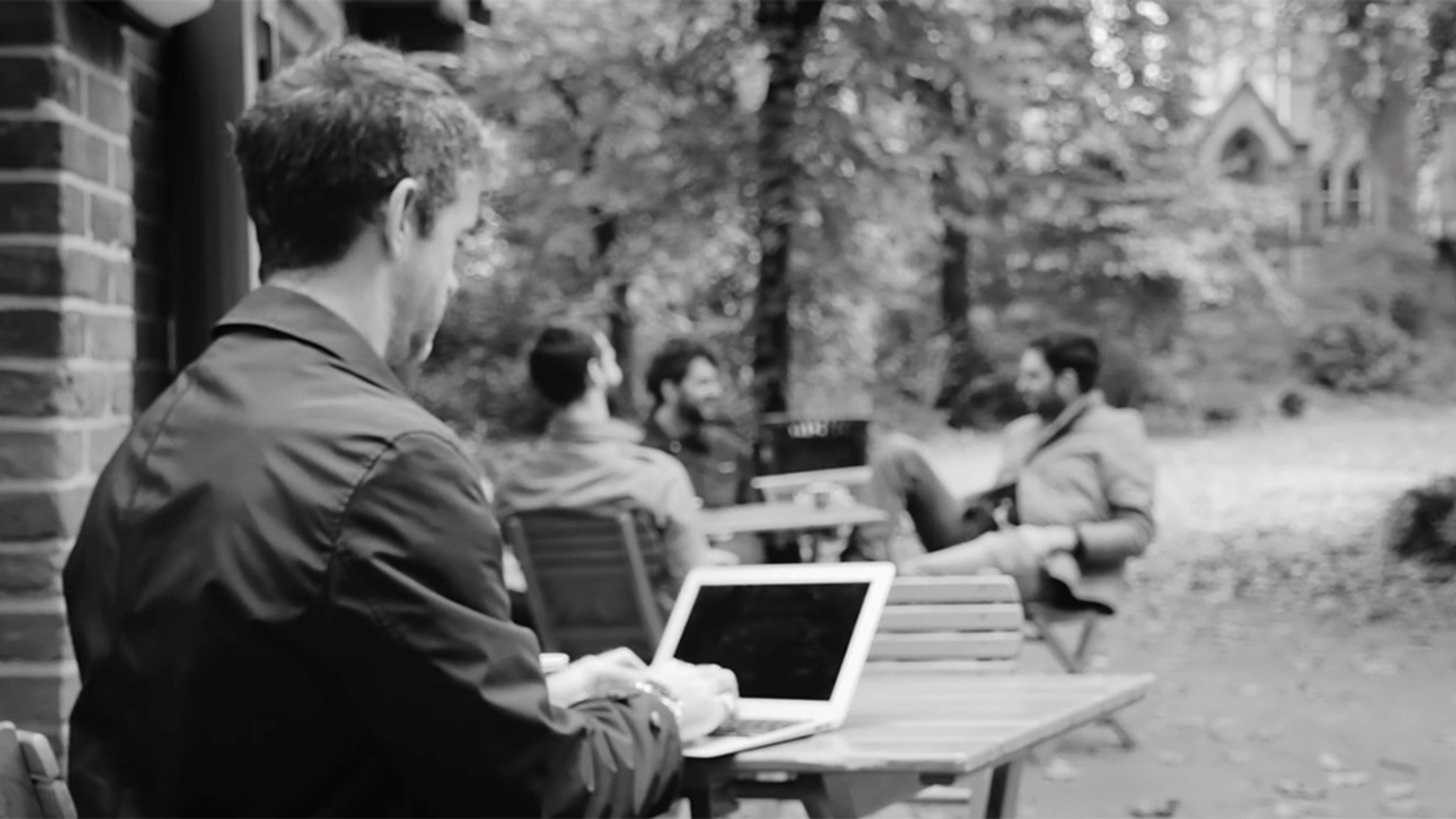 Black and white photo of a man working on a laptop outdoors with three men talking in the background in a wooded area.