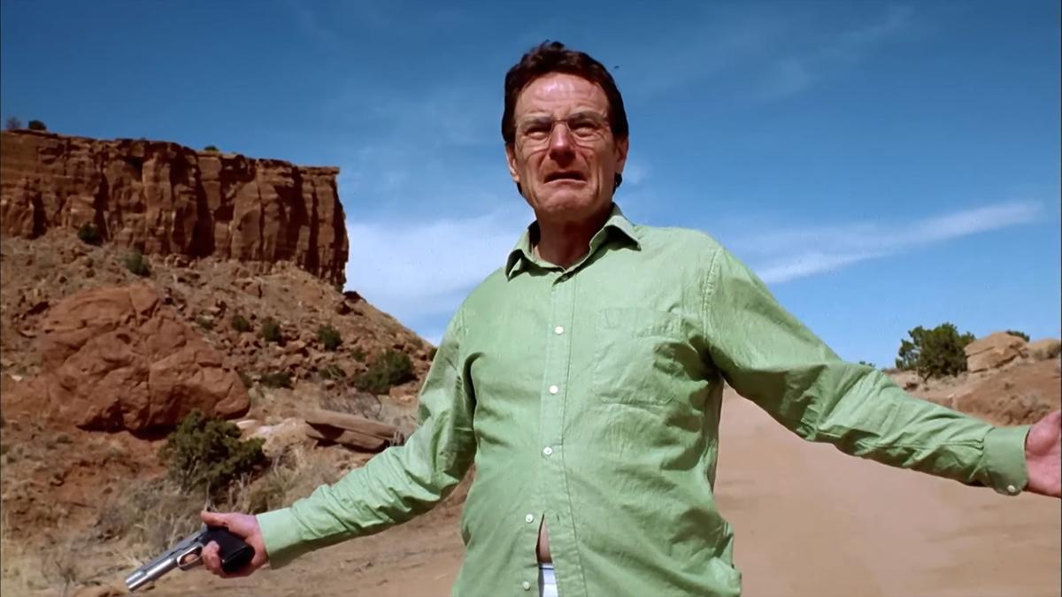 Photo of a man in a green shirt holding a gun in a desert landscape with rocky cliffs under a blue sky.
