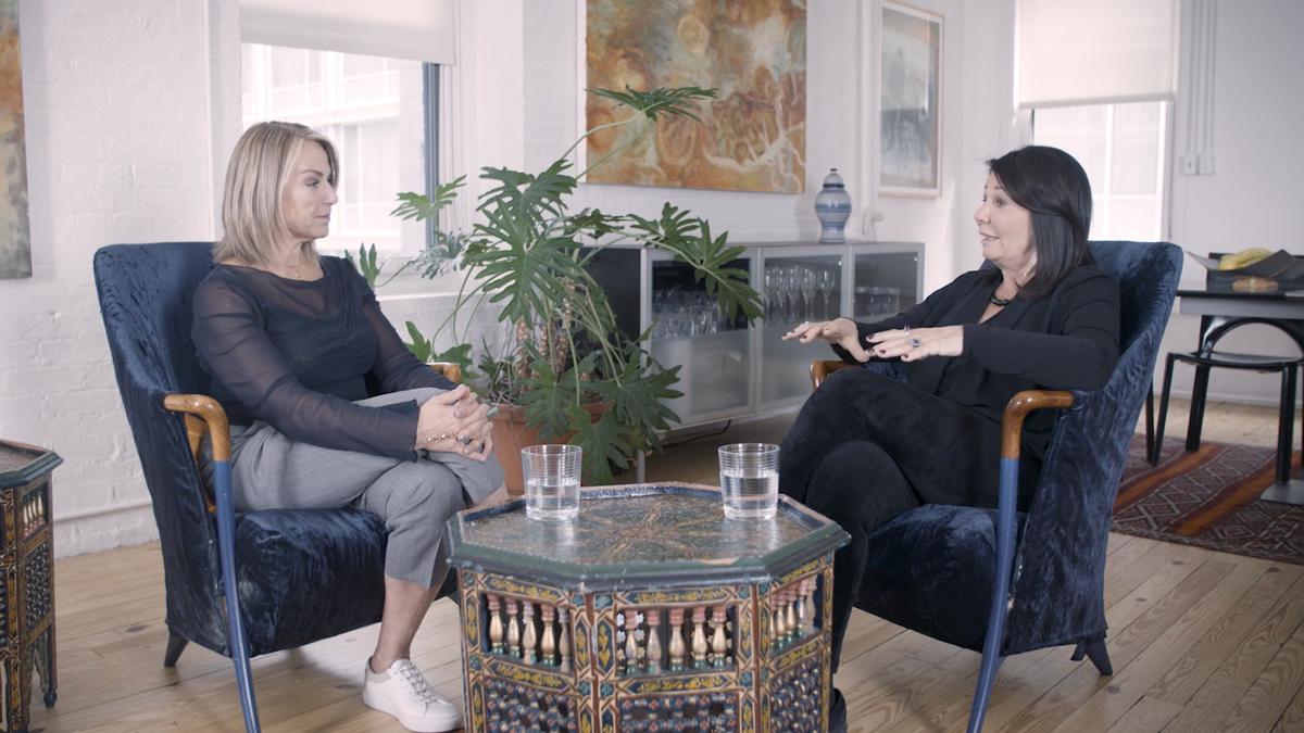 Photo of two women sitting in blue chairs talking in a bright room with plants, a patterned table between them.