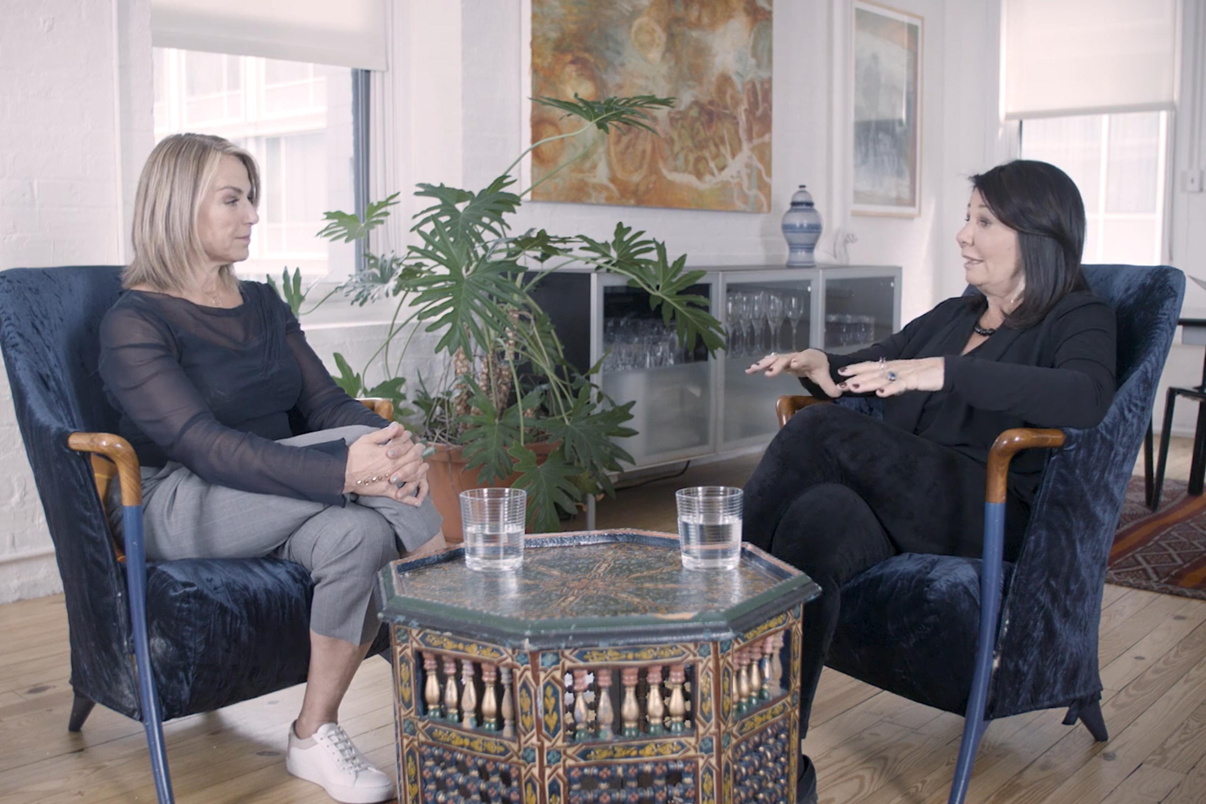 Photo of two women sitting in blue chairs talking in a bright room with plants, a patterned table between them.