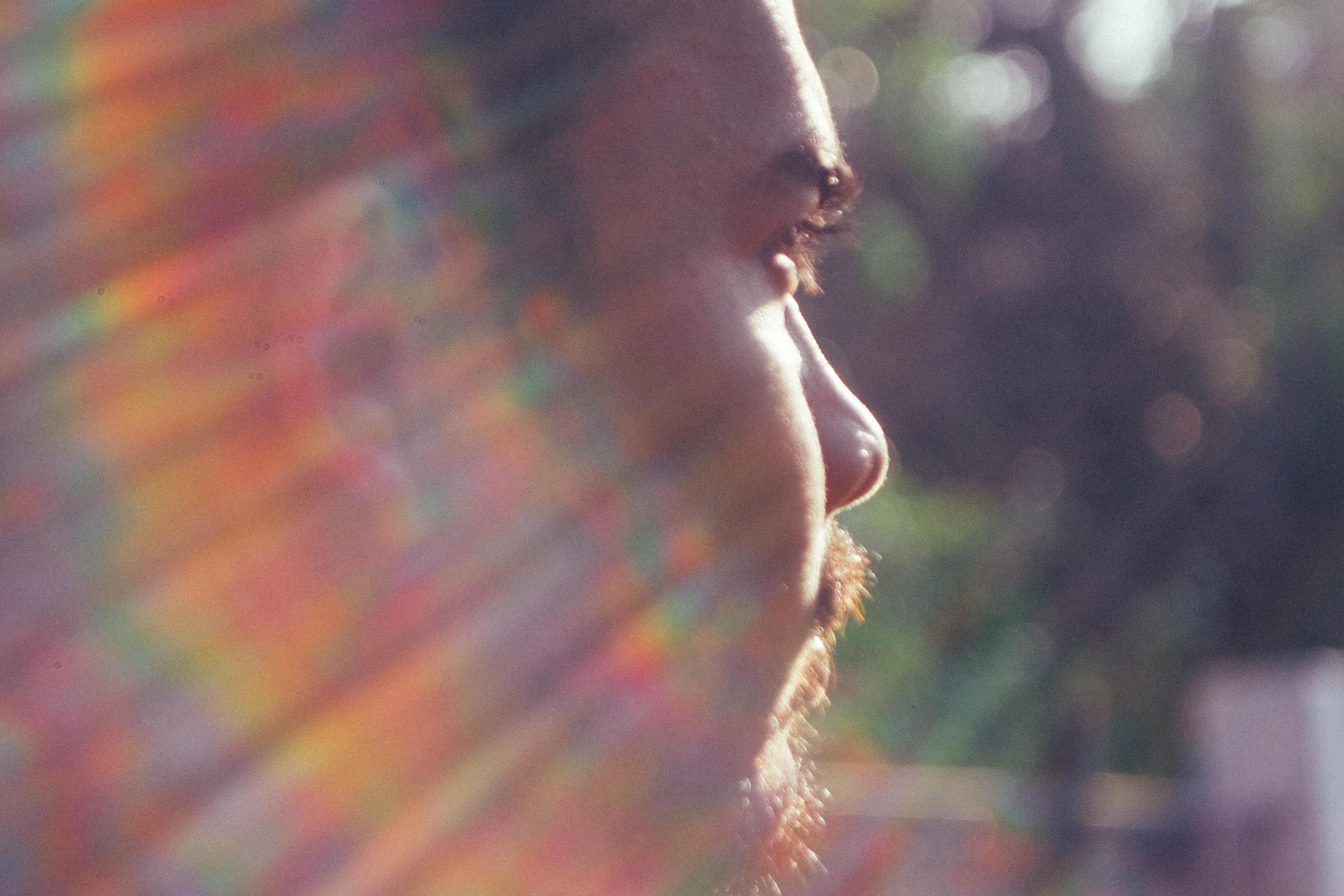 Photo of a man’s side profile with sunlight creating rainbow flares across his face, blurred greenery background.
