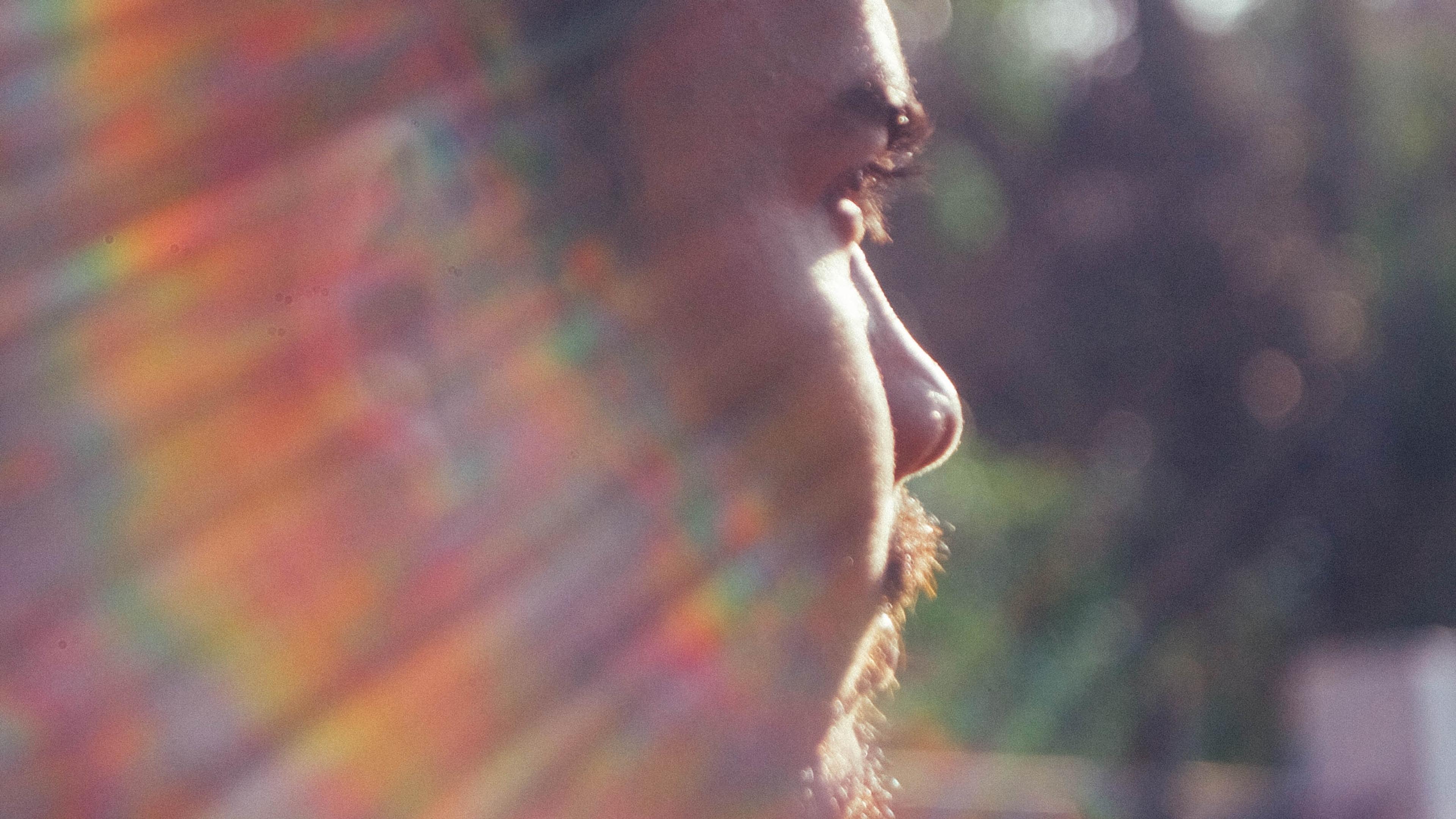 Photo of a man’s side profile with sunlight creating rainbow flares across his face, blurred greenery background.