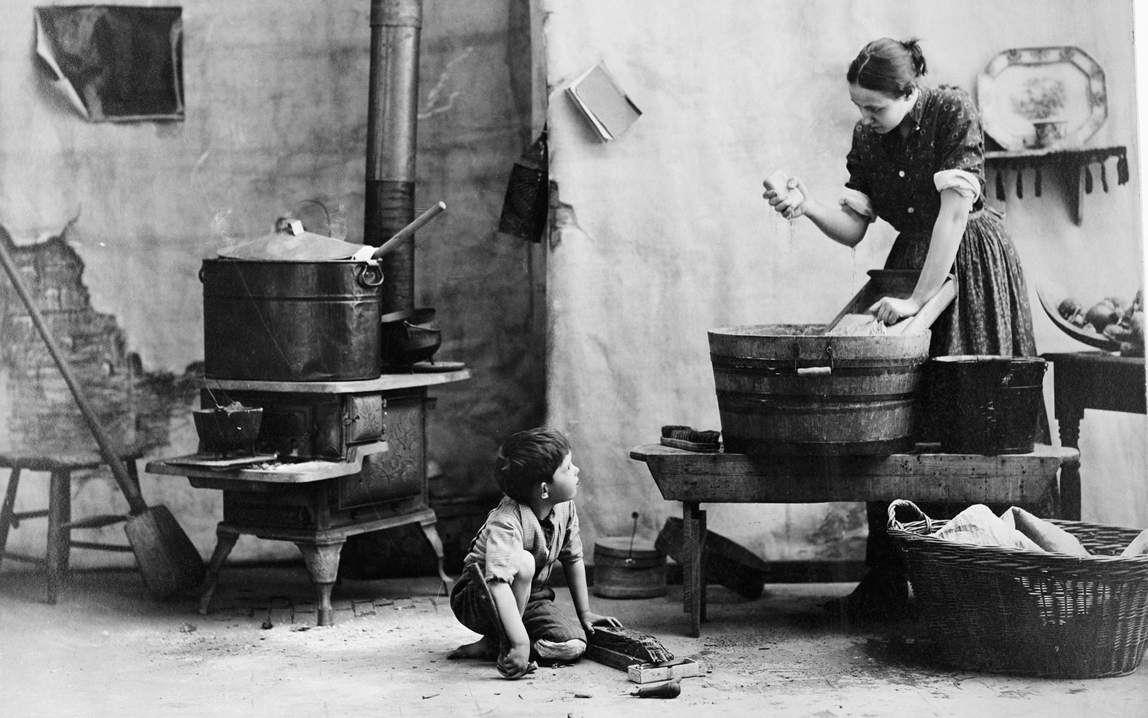 Vintage photo of a woman doing laundry in a wooden tub while a child watches, set in a rustic room with a stove.