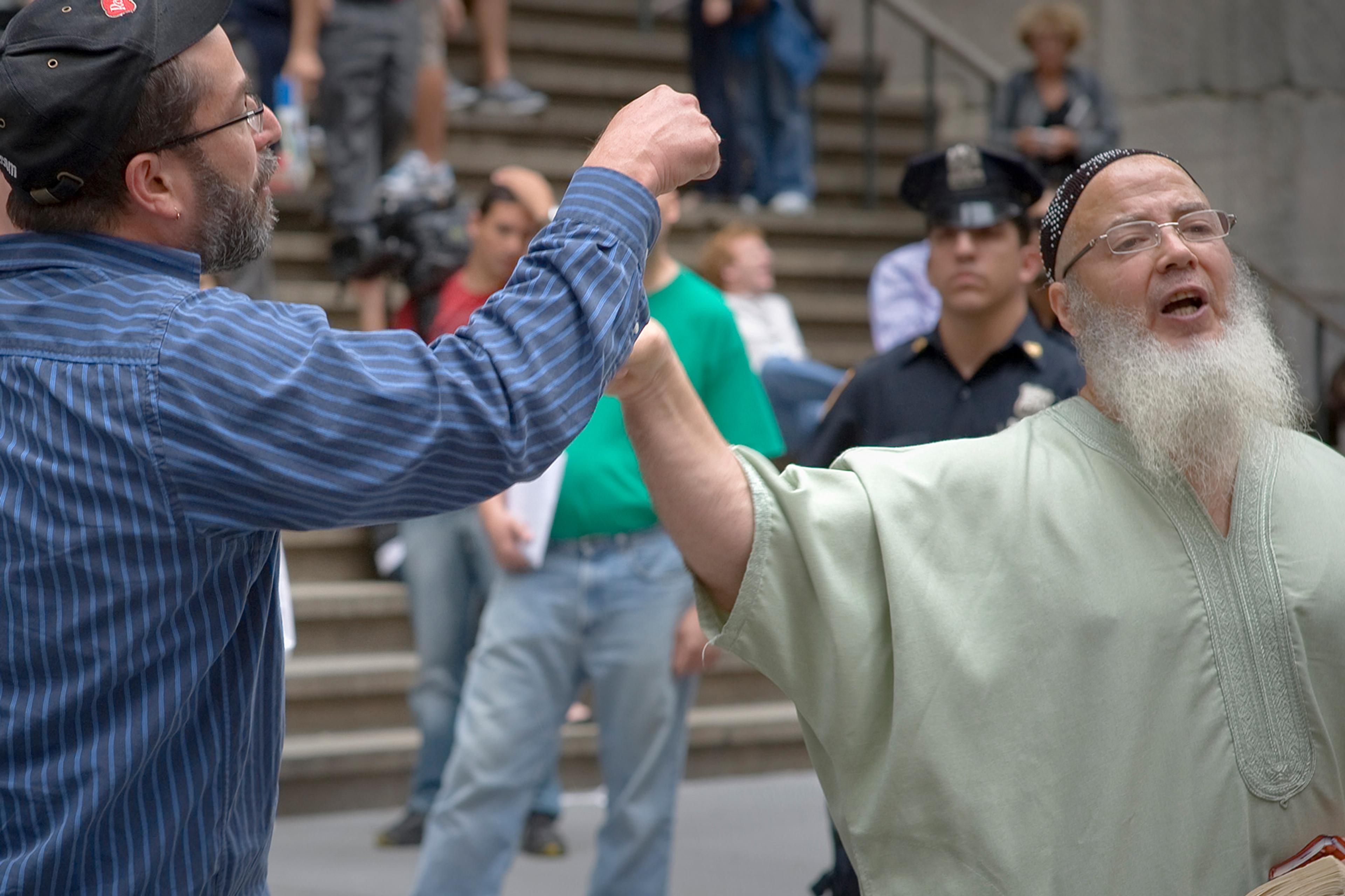 A man in European clothing and a man in a skullcap, loose robe and white beard gesticulate while a police officer stands in the background.