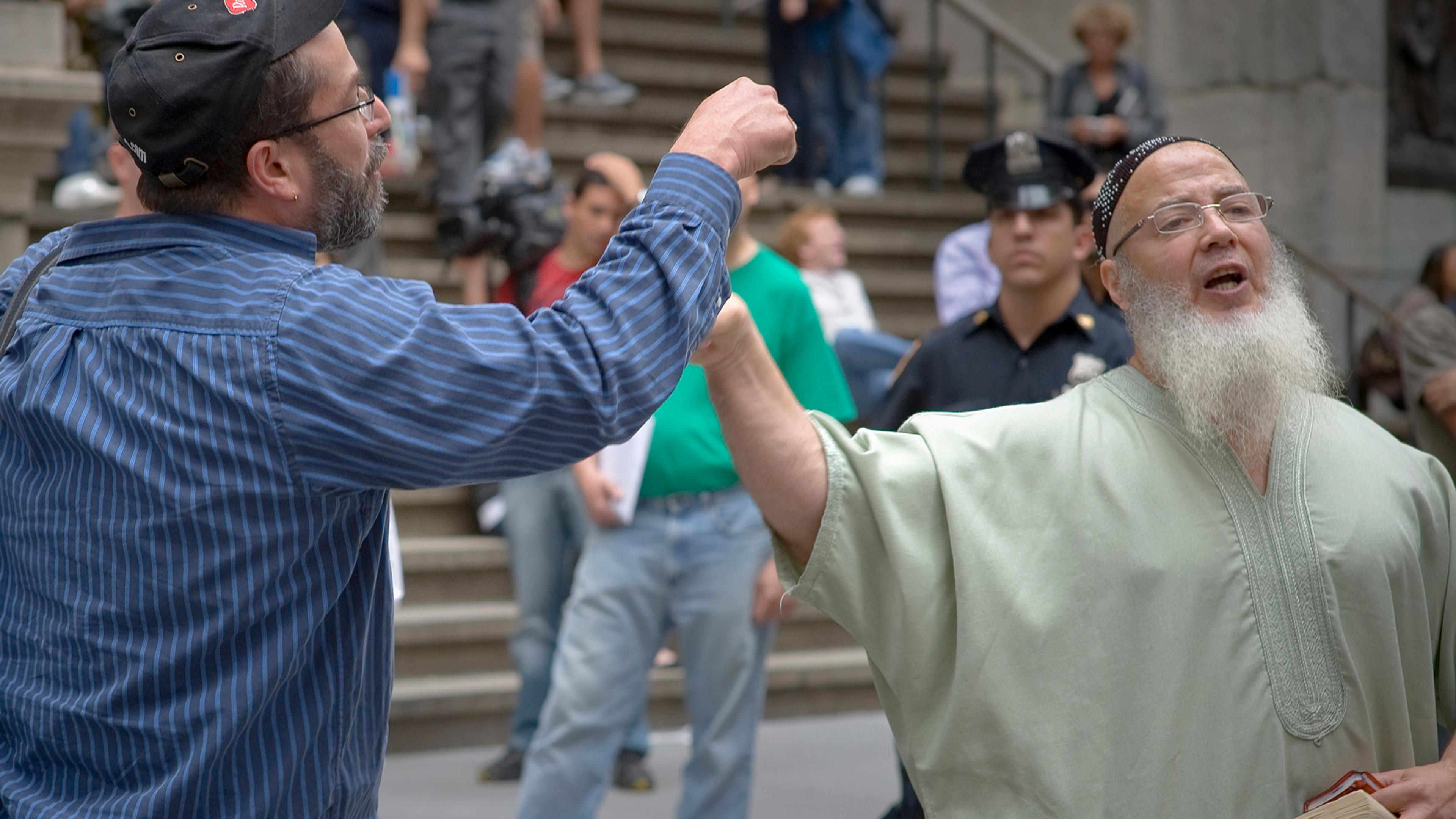 A man in European clothing and a man in a skullcap, loose robe and white beard gesticulate while a police officer stands in the background.