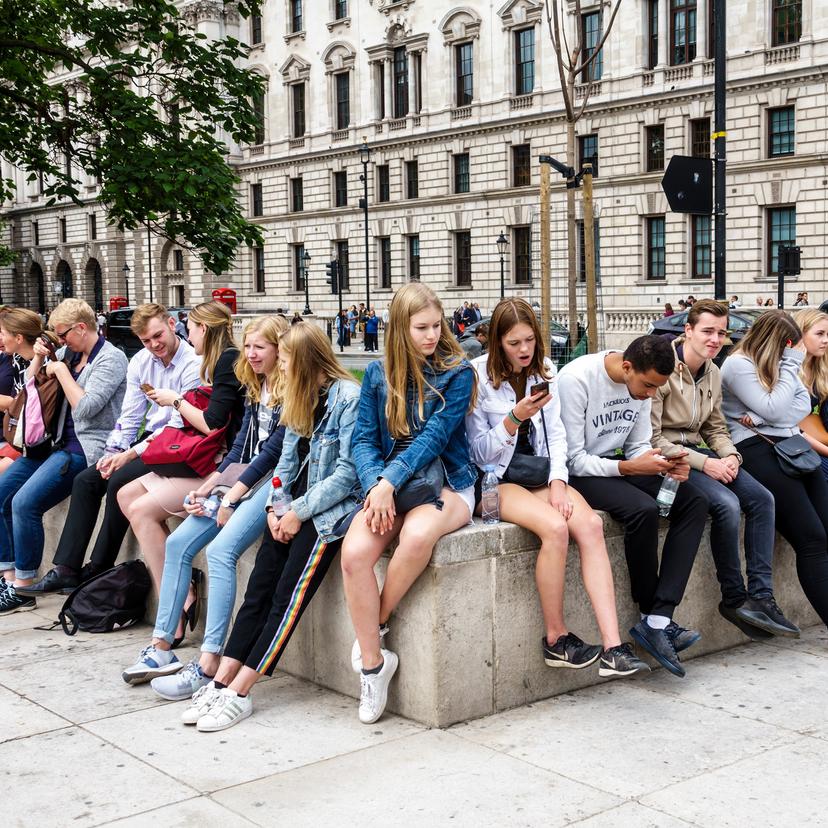 Photo of a group of young people in casual clothing sitting on a concrete bench outside a historic building, some using phones.