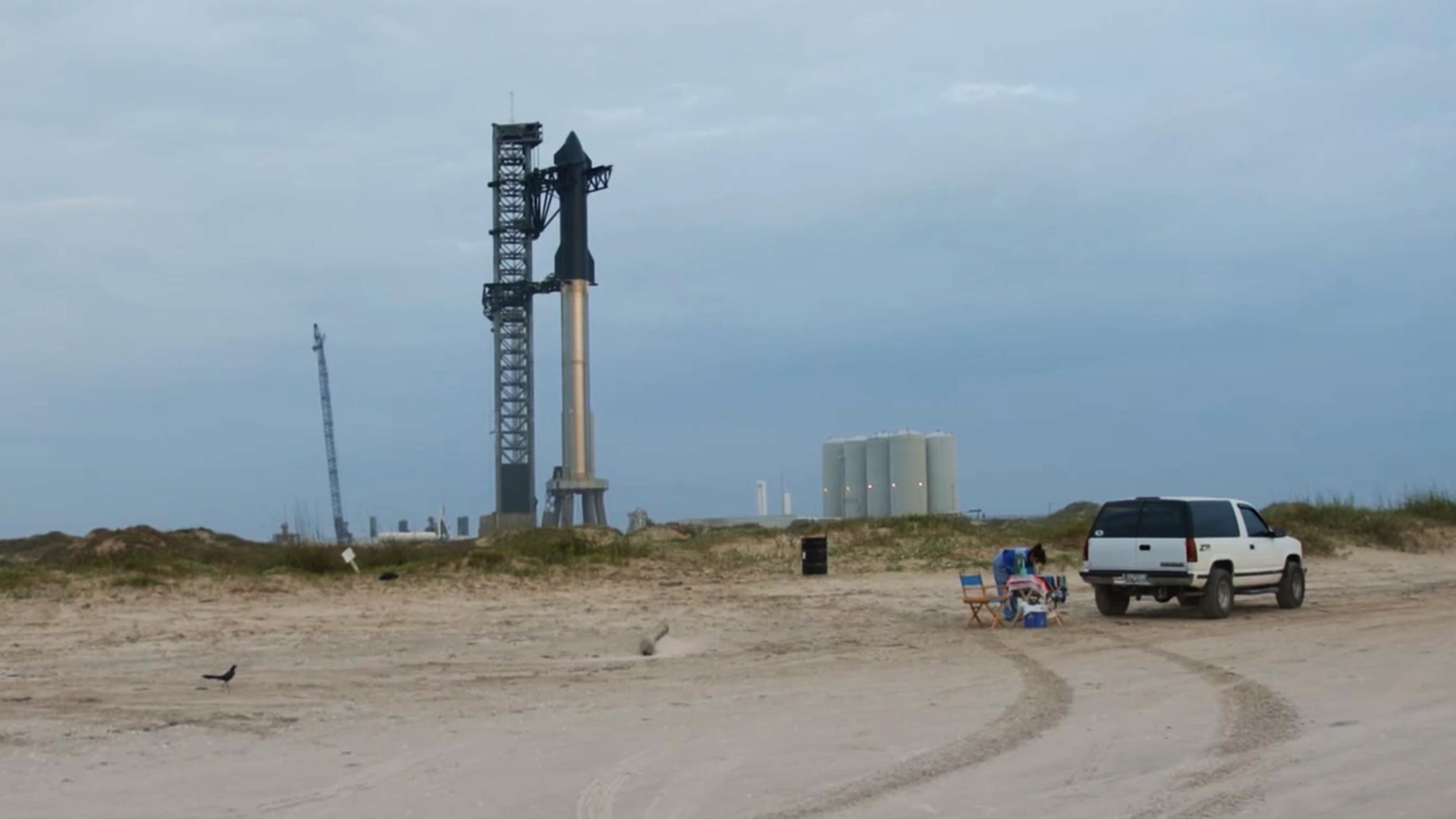 A beach with a white S U V, chairs and a bird in the foreground, and a rocket on a launch pad in the background.