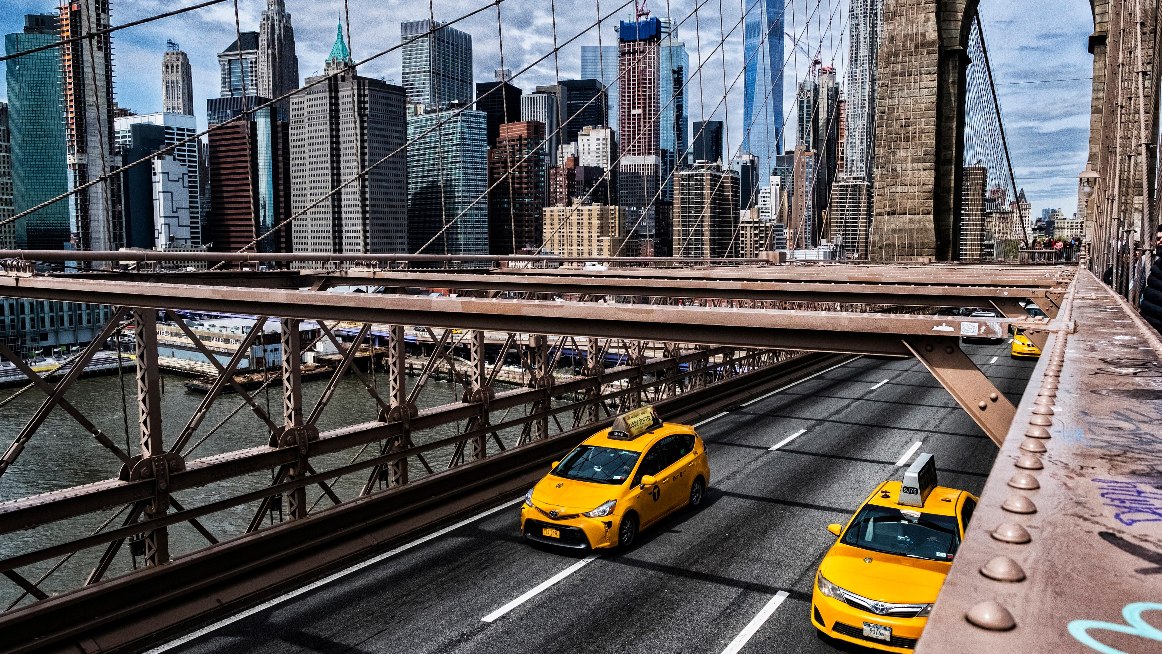 Photo of yellow taxis on Brooklyn Bridge with New York City skyline in the background on a cloudy day.