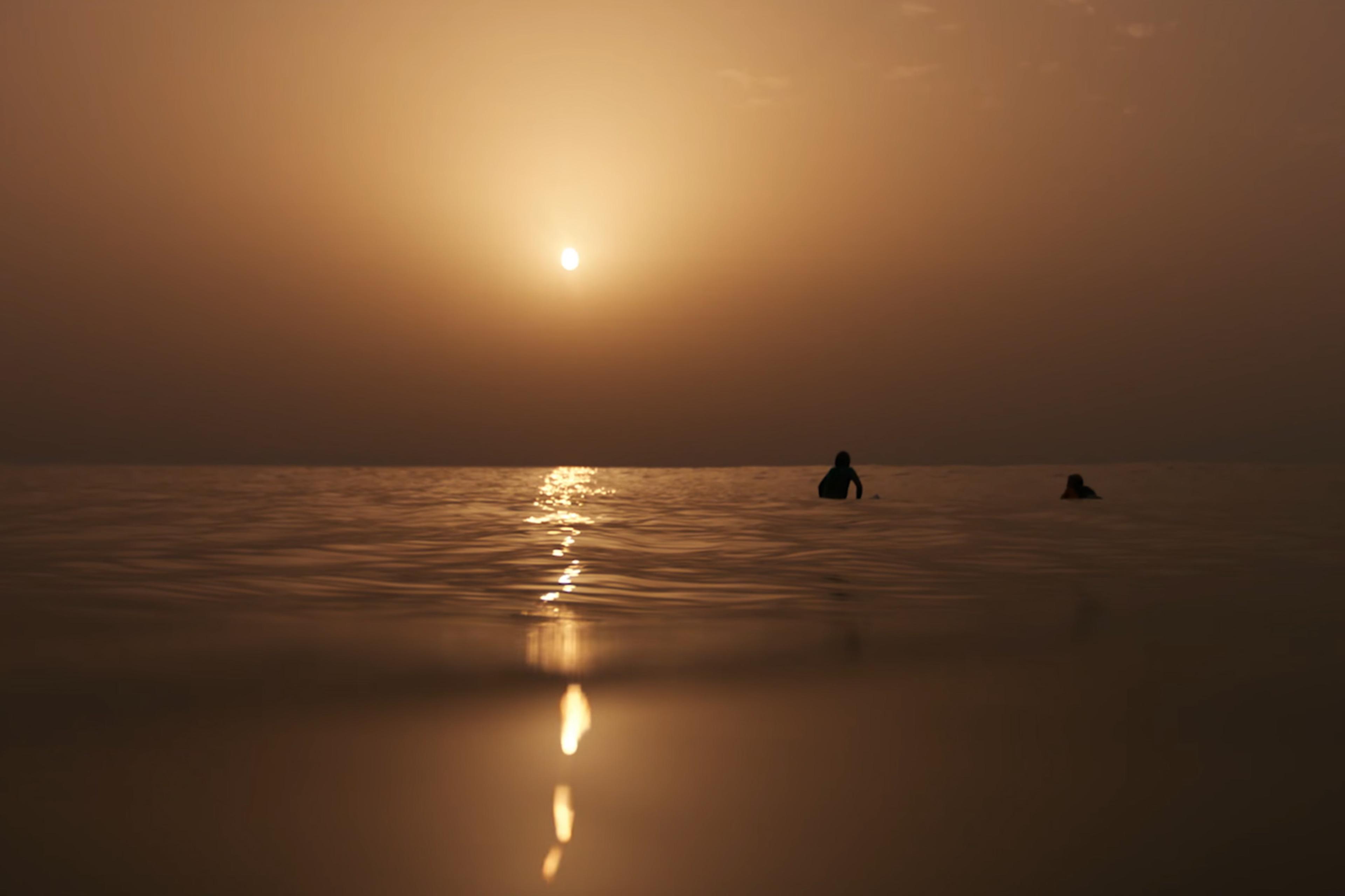 A sunset over the sea with two people swimming. The sun’s reflection casts a golden glow on the calm water.