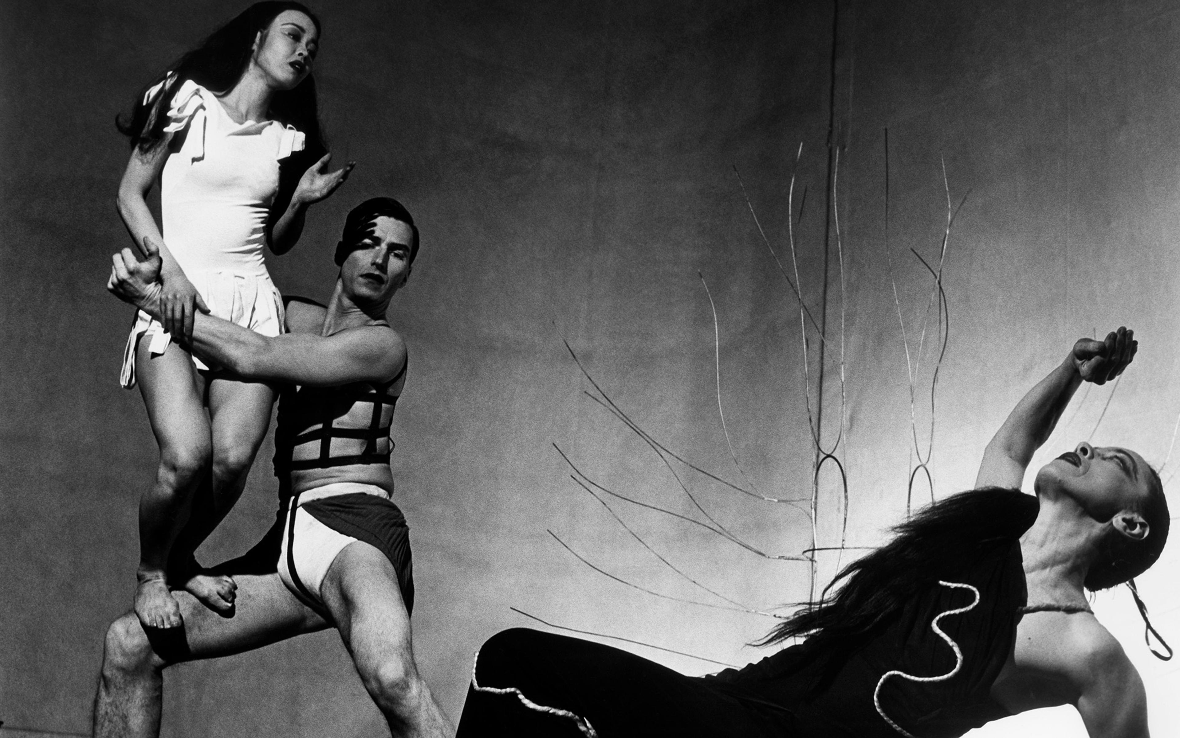 Black-and-white photo of three dancers in dynamic poses; one man lifting a woman in a dress while another woman in a black dress dramatically leans back.