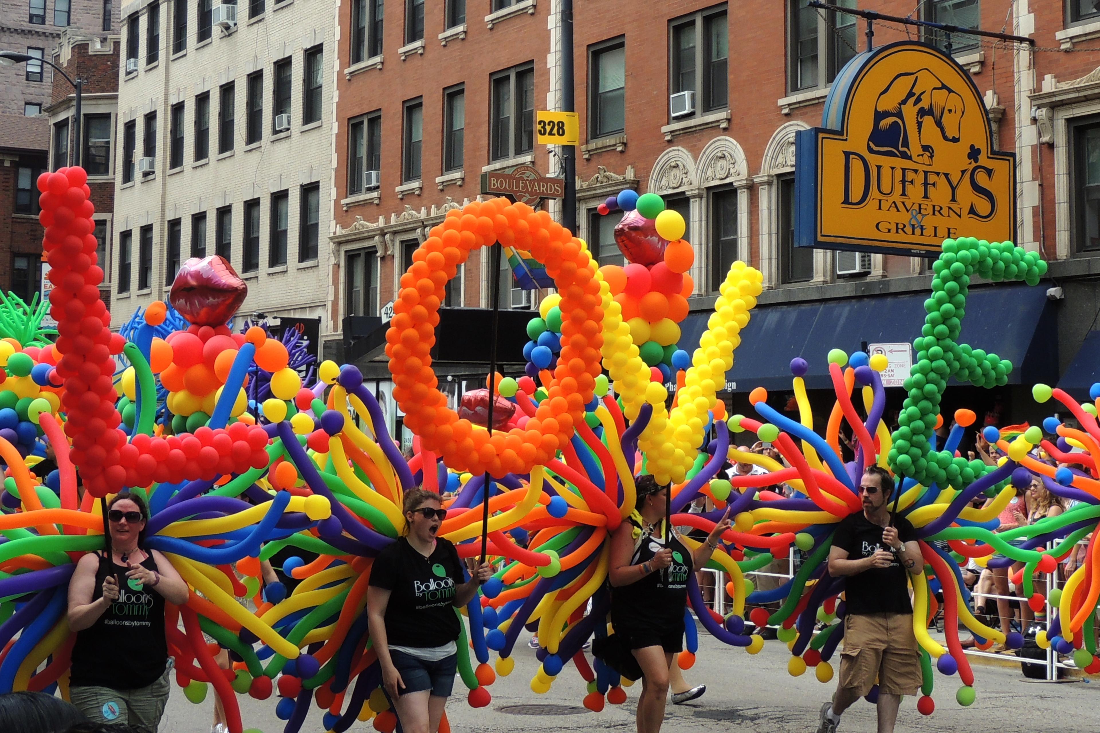 Photo of a colourful parade float spelling “LOVE” with balloons in an urban street setting.