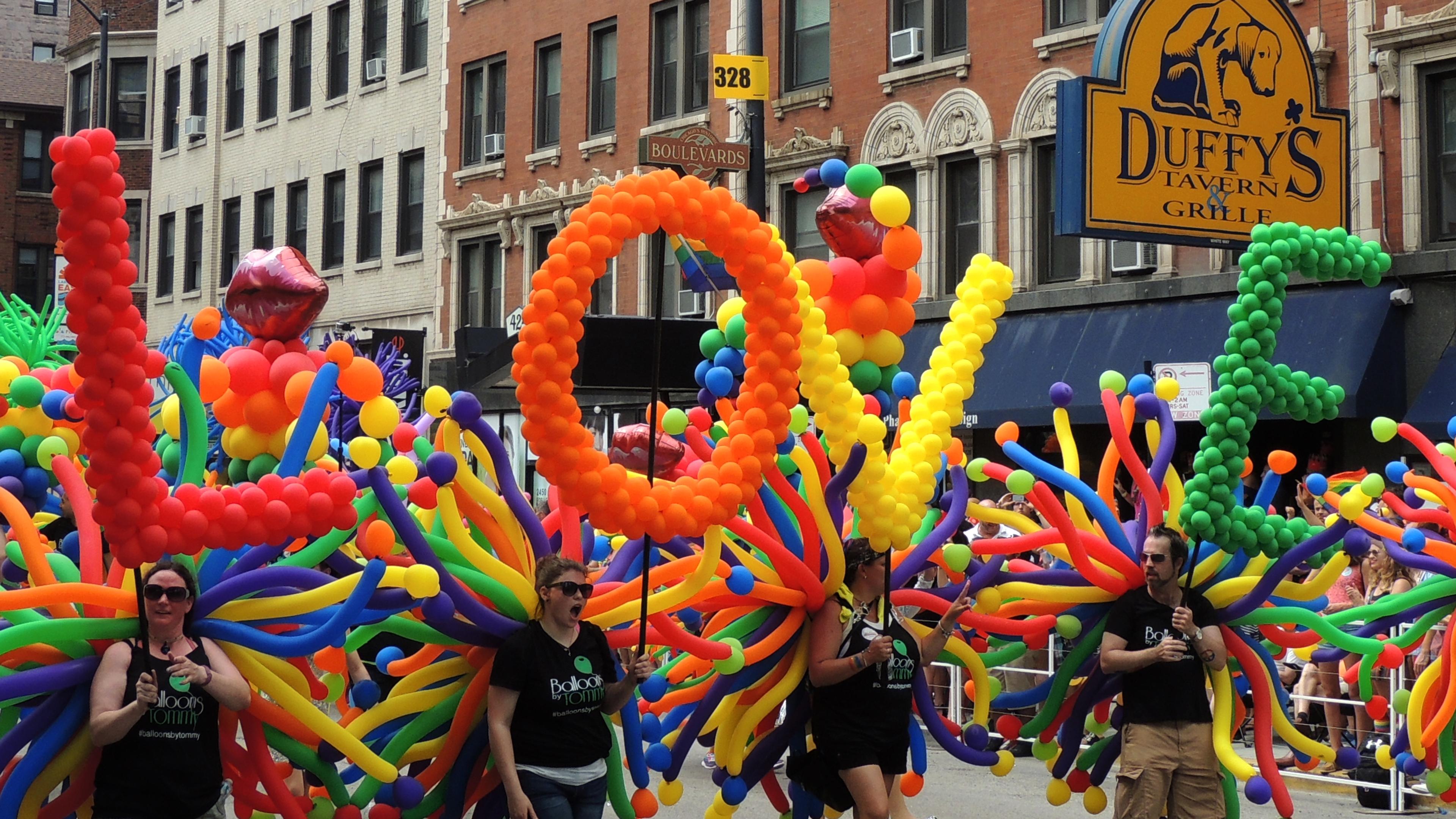 Photo of a colourful parade float spelling “LOVE” with balloons in an urban street setting.
