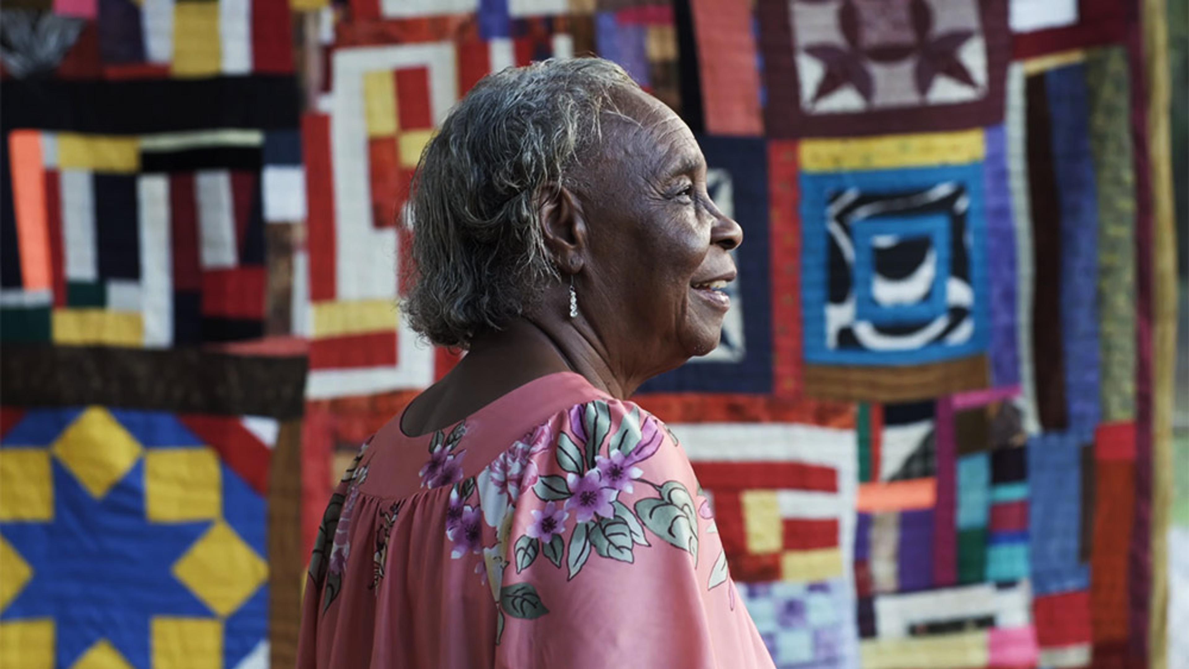 An elderly Black woman in a pink floral dress standing in front of colourful quilts with various geometric patterns.