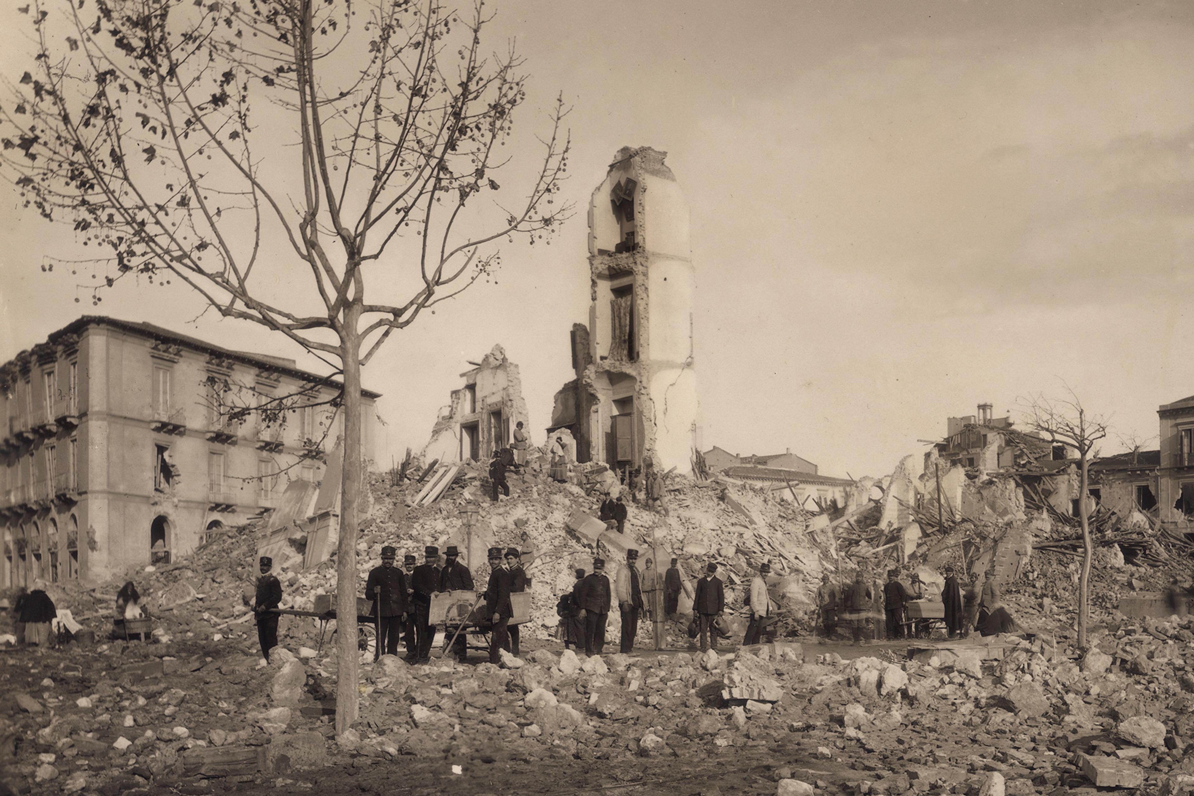 Historic photo of a group gathered near ruins of a building after destruction. A leafless tree stands in foreground.