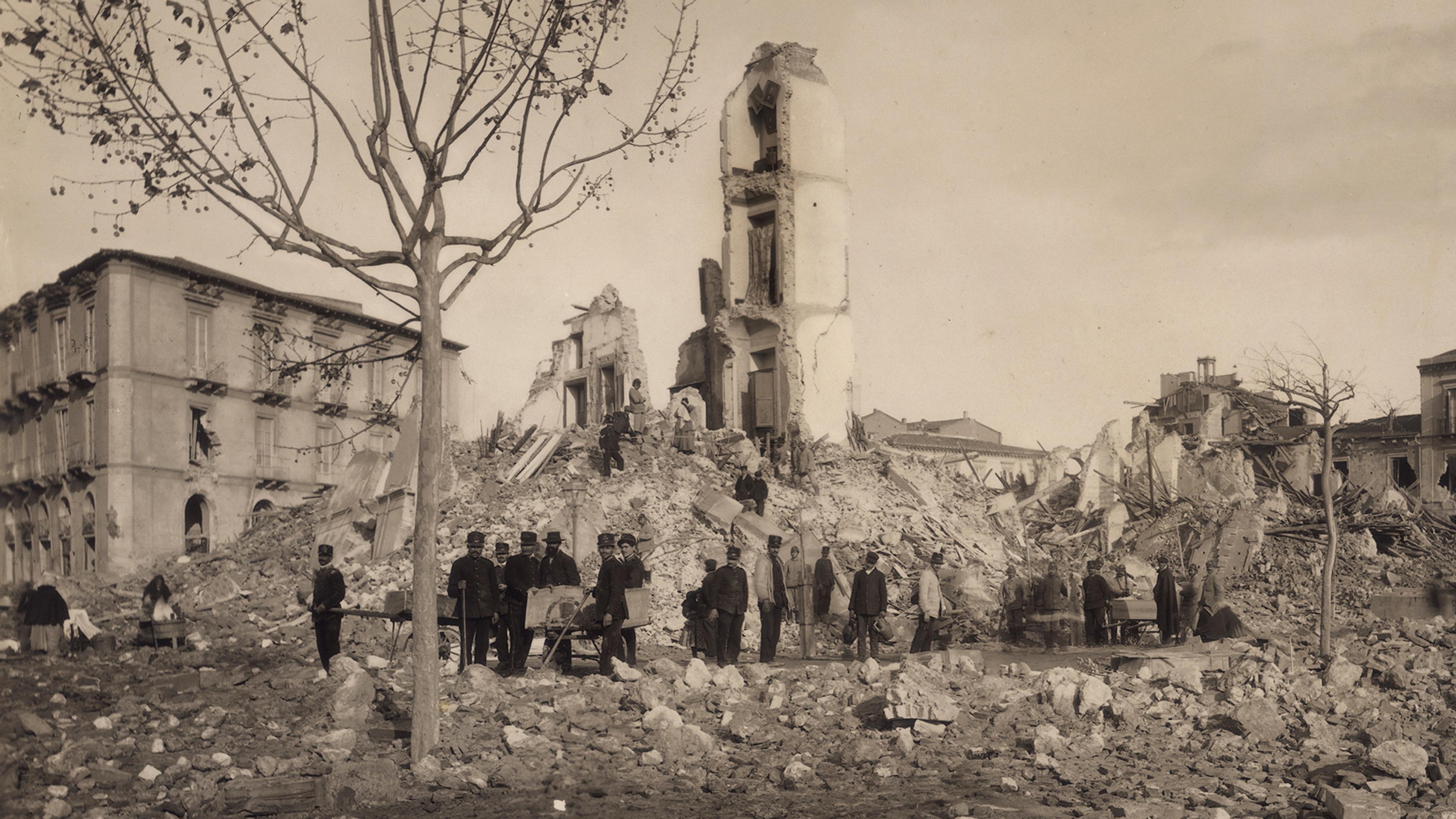 Historic photo of a group gathered near ruins of a building after destruction. A leafless tree stands in foreground.