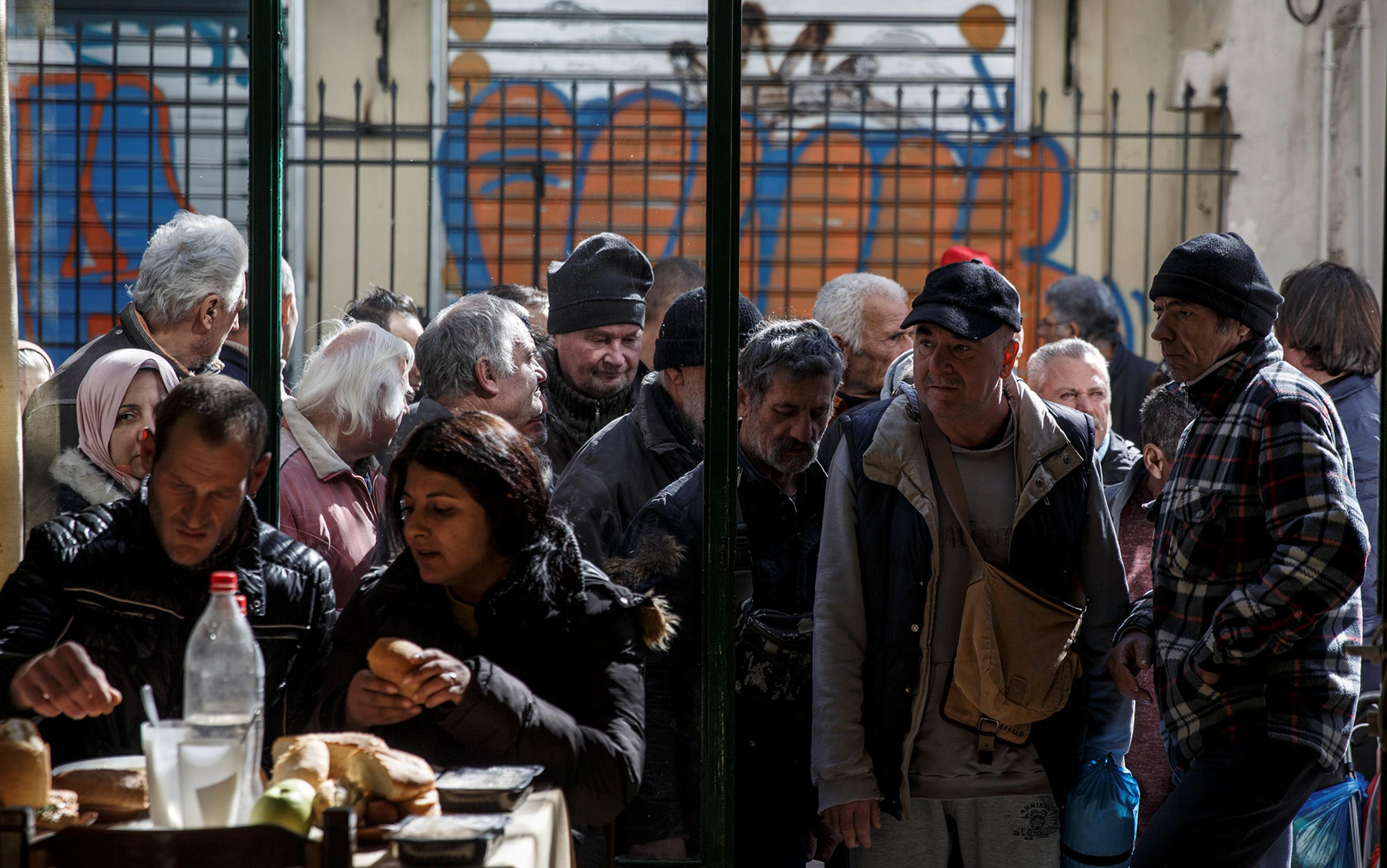 Seen from inside, a couple eat a basic meal whilst outside in an alleyway a queue of mostly older people wait their turn