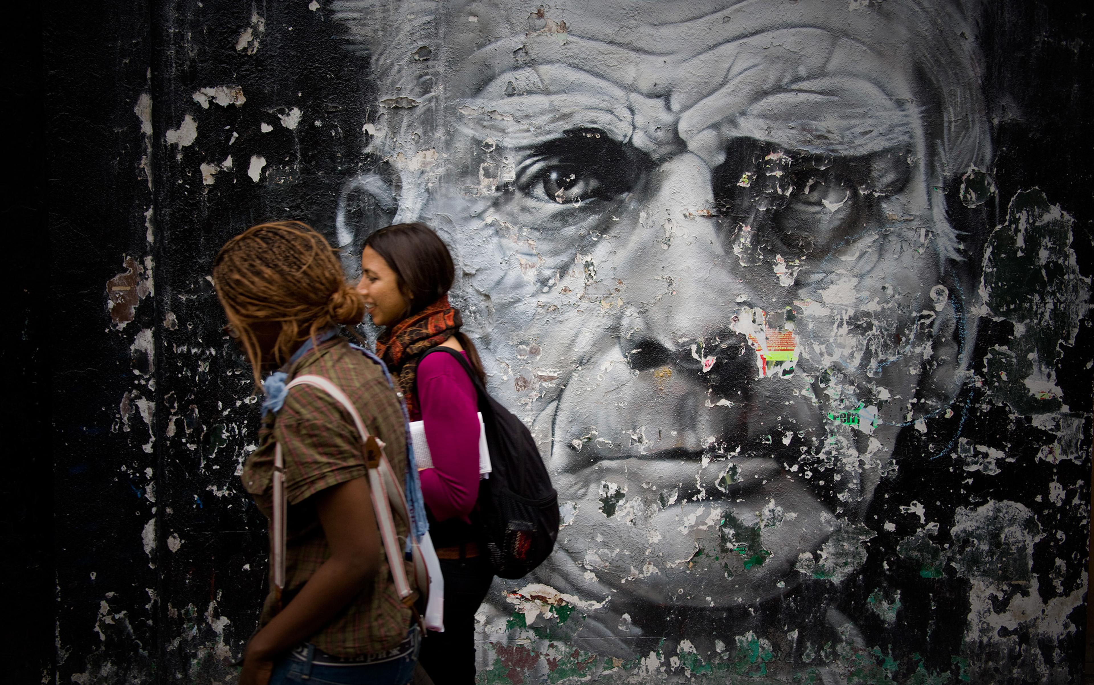 Two young women walking past a weathered mural of Samuel Beckett’s face on a wall with peeling paint and worn textures.