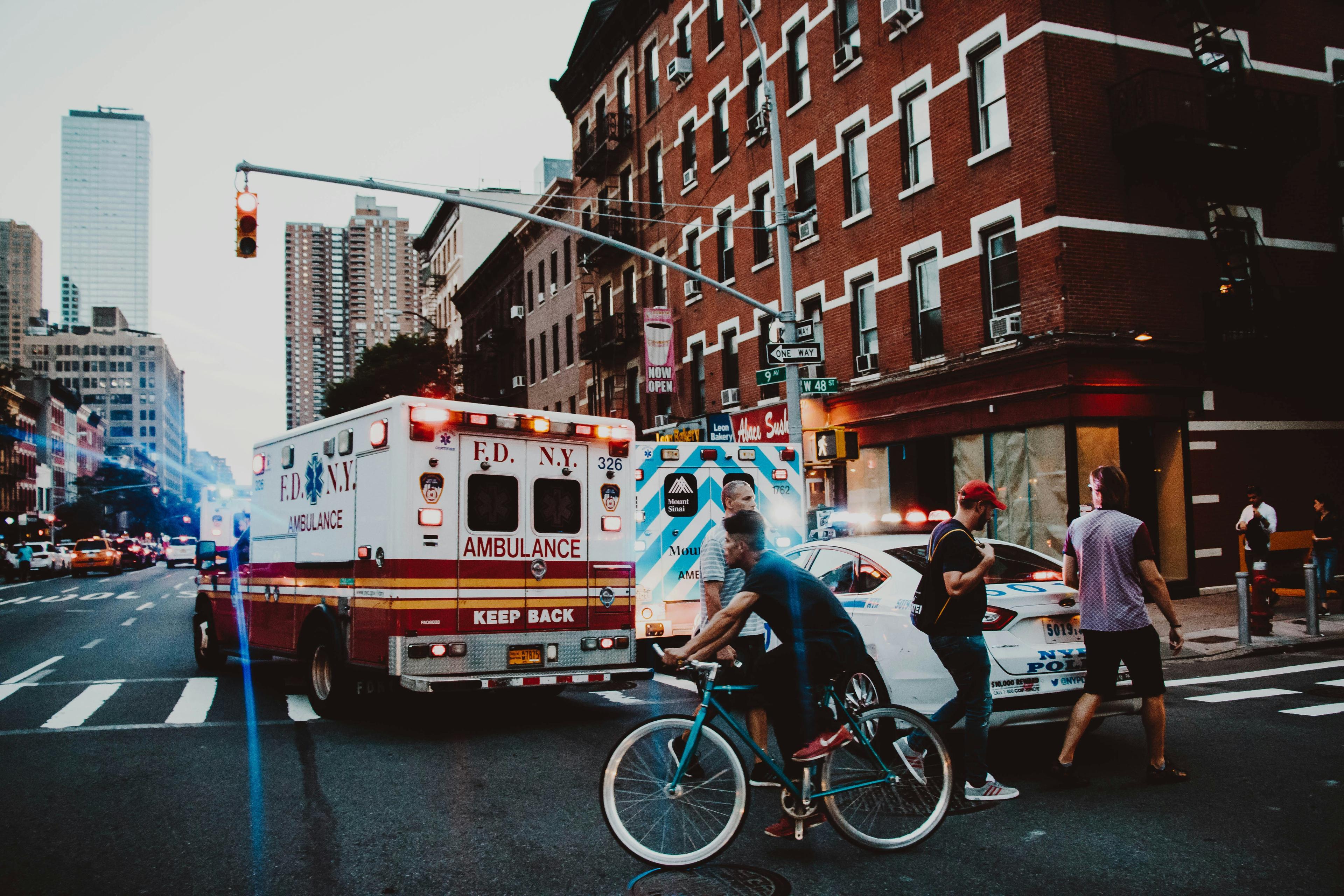 A busy city street with ambulances and a police car. People walk and cycle nearby under tall buildings and traffic lights.