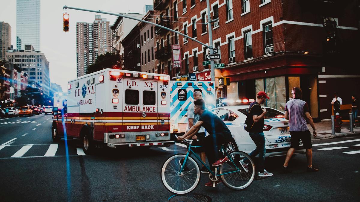 A busy city street with ambulances and a police car. People walk and cycle nearby under tall buildings and traffic lights.