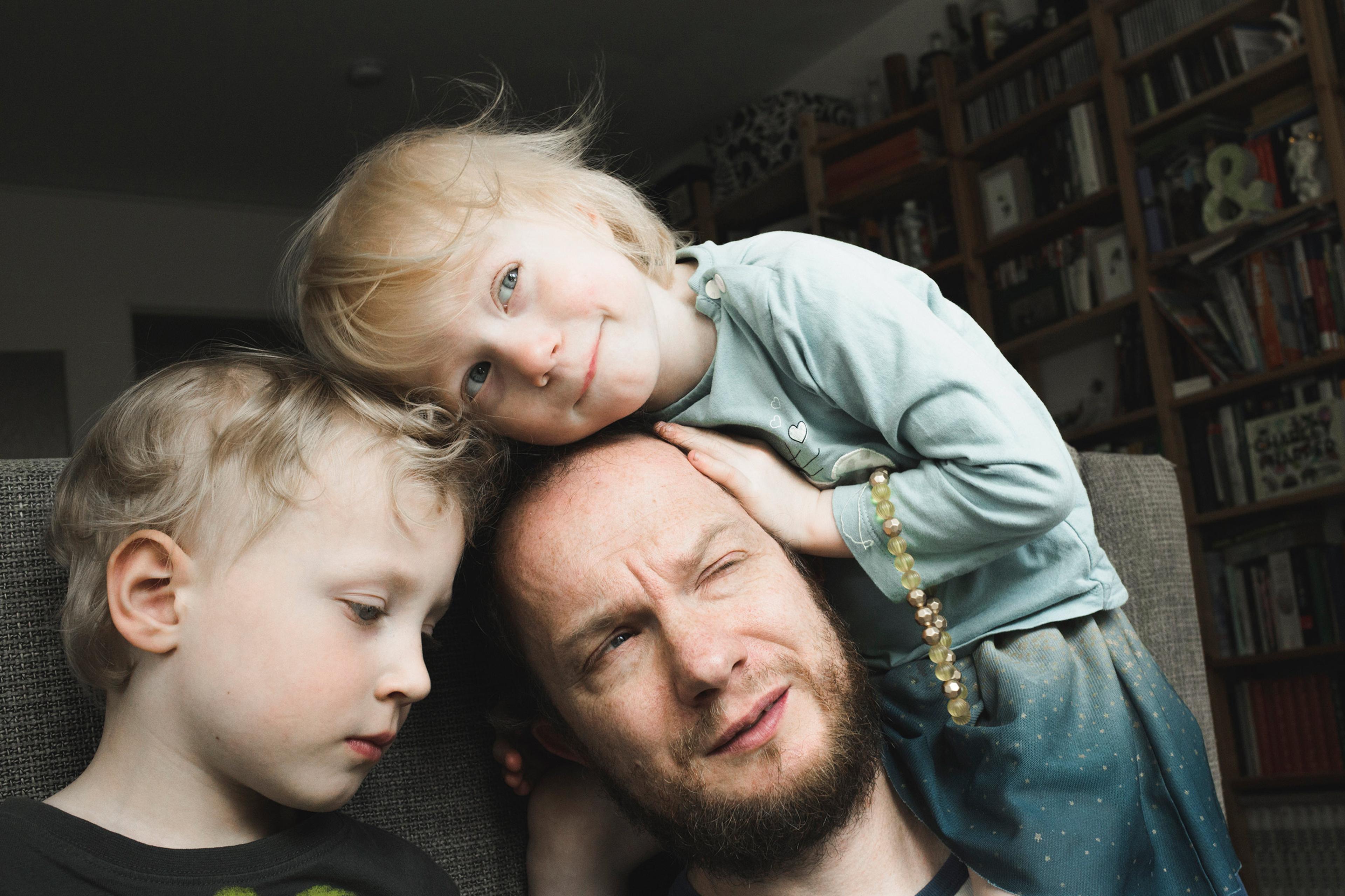 A man with a beard sitting on a sofa with two young children, one resting on his head, in a room with bookshelves.
