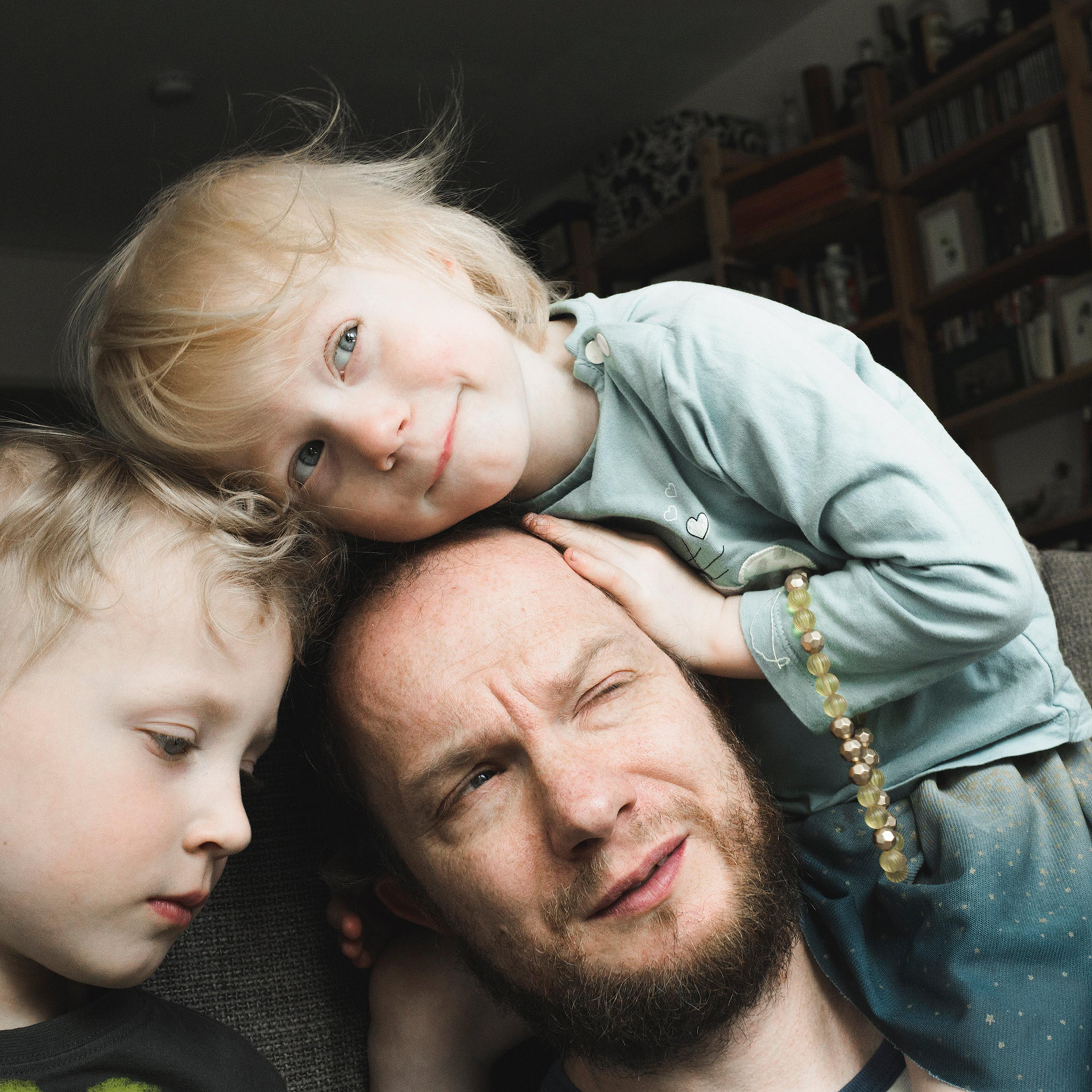 A man with a beard sitting on a sofa with two young children, one resting on his head, in a room with bookshelves.
