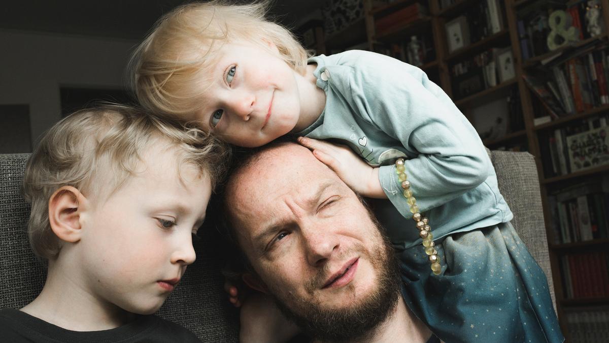 A man with a beard sitting on a sofa with two young children, one resting on his head, in a room with bookshelves.