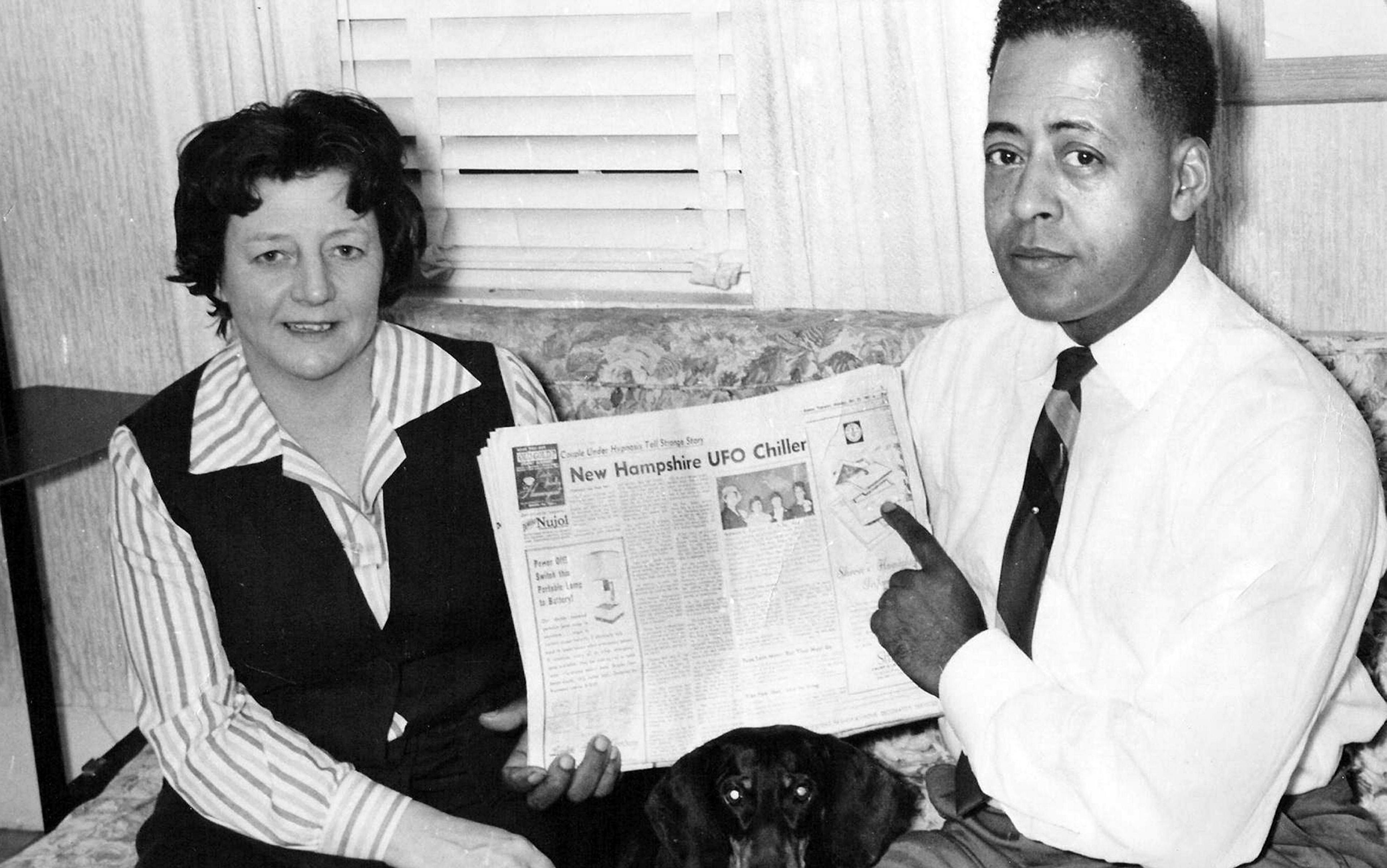 Black and white photo of a woman and man sitting on a sofa holding a newspaper with a front-page story about a UFO sighting.