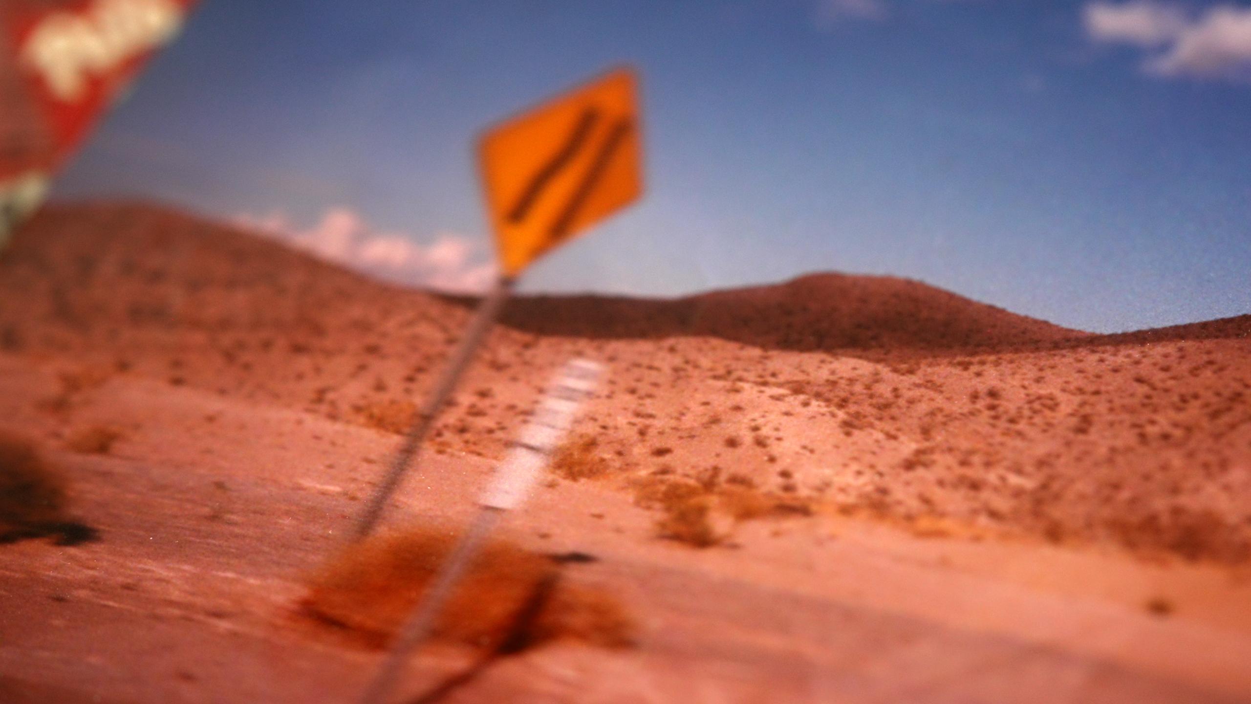 Desert with a blurred yellow road sign and distant hills under a blue sky.