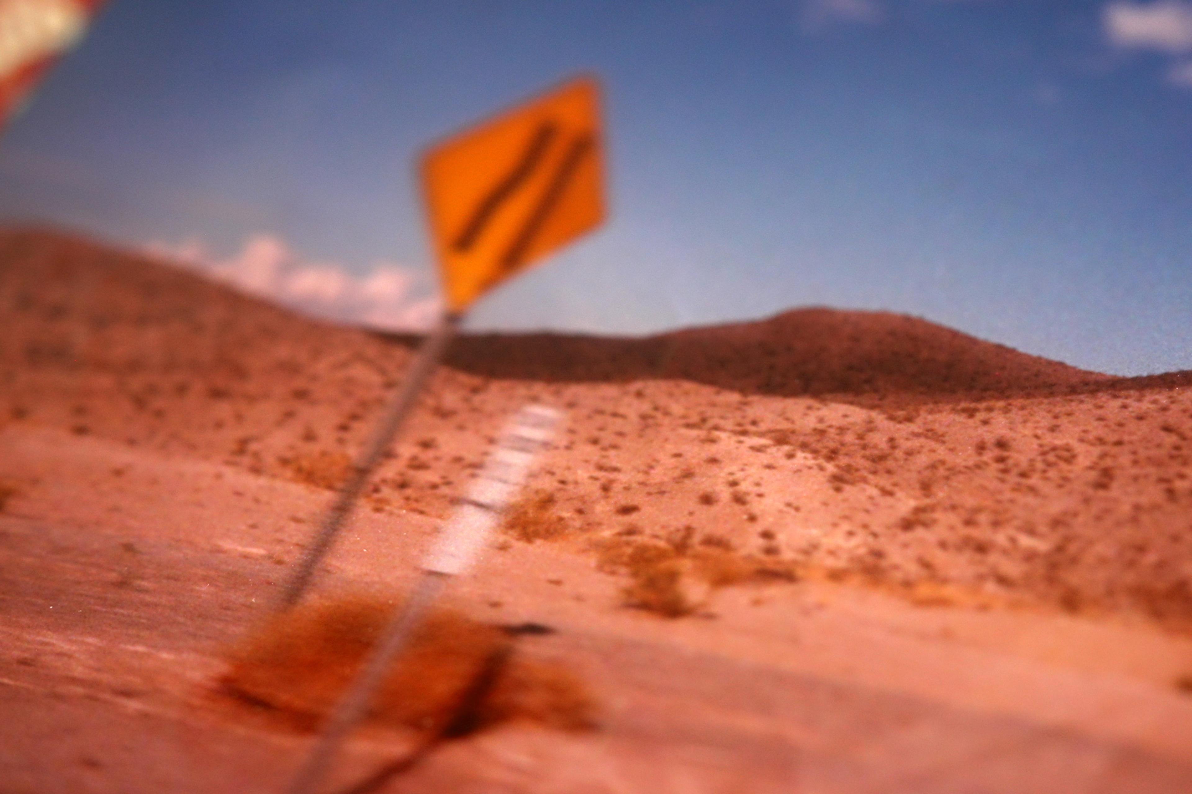 Desert with a blurred yellow road sign and distant hills under a blue sky.