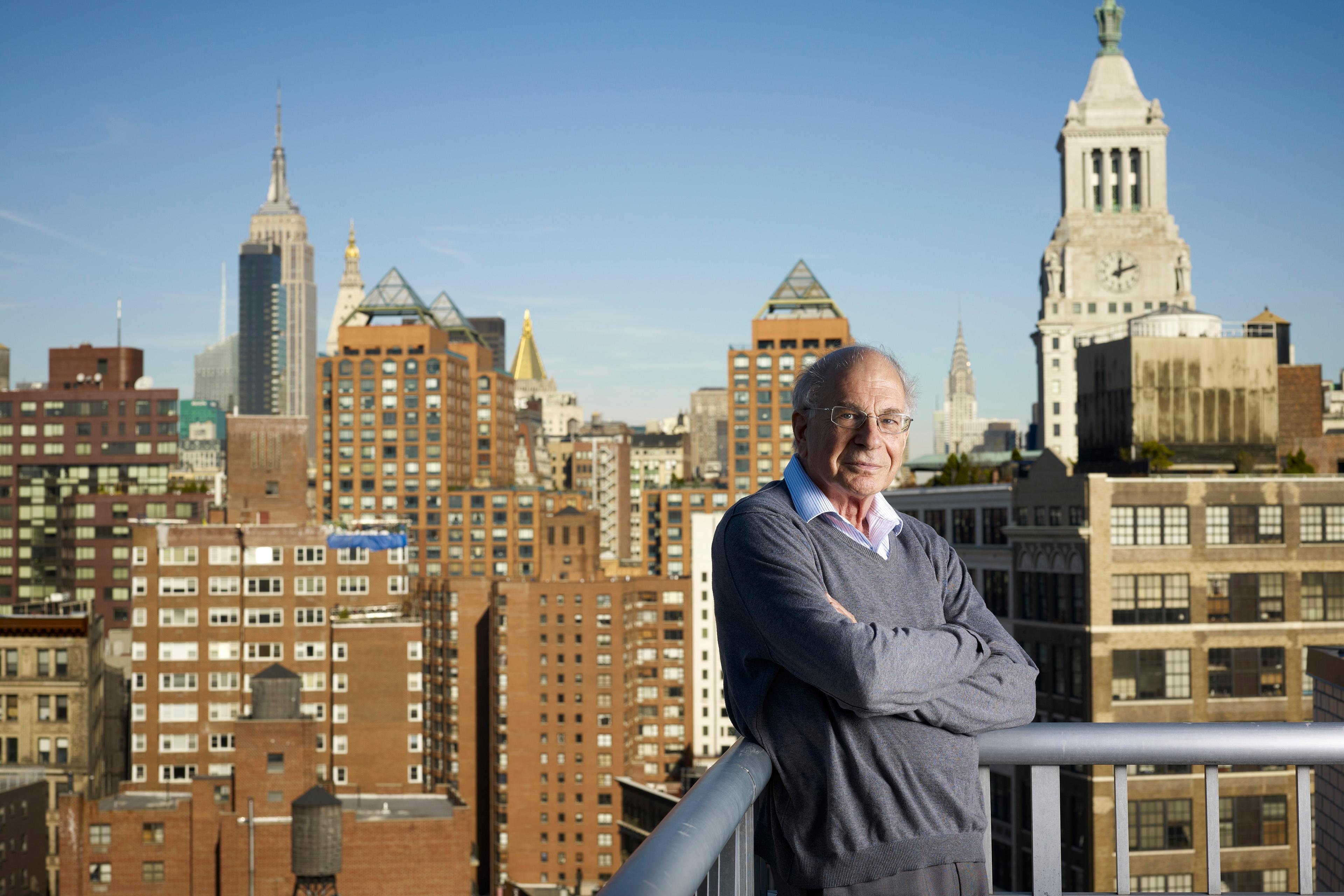 Photo of a man in a grey jumper standing on a balcony with a view of a city skyline featuring iconic skyscrapers.