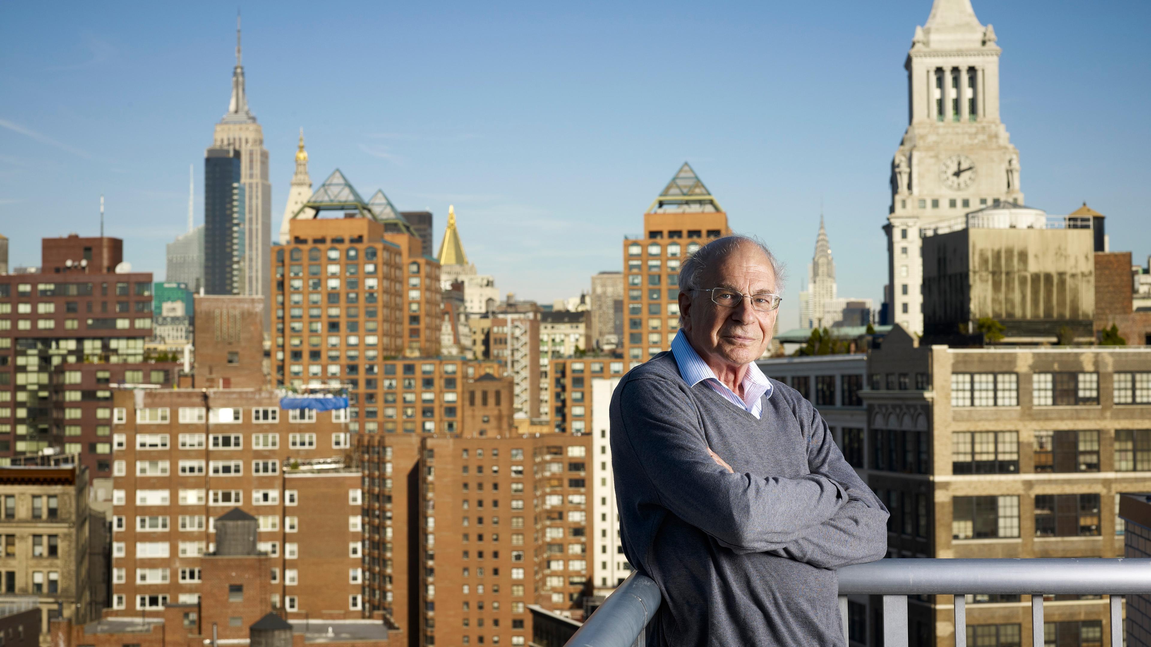 Photo of a man in a grey jumper standing on a balcony with a view of a city skyline featuring iconic skyscrapers.