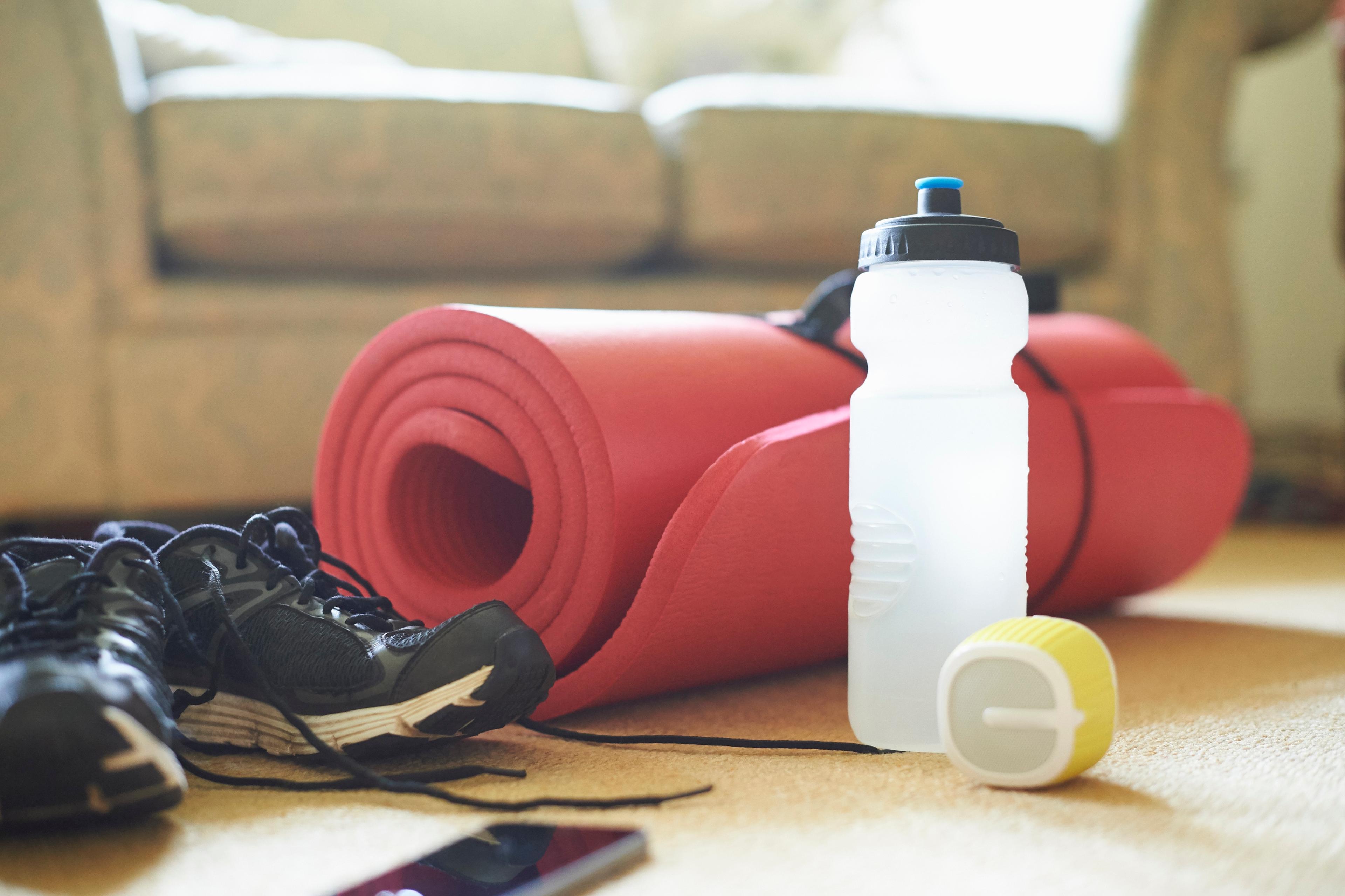 Photo of gym shoes, a red rolled yoga mat, a water bottle and a speaker on a carpeted floor in a cosy room.