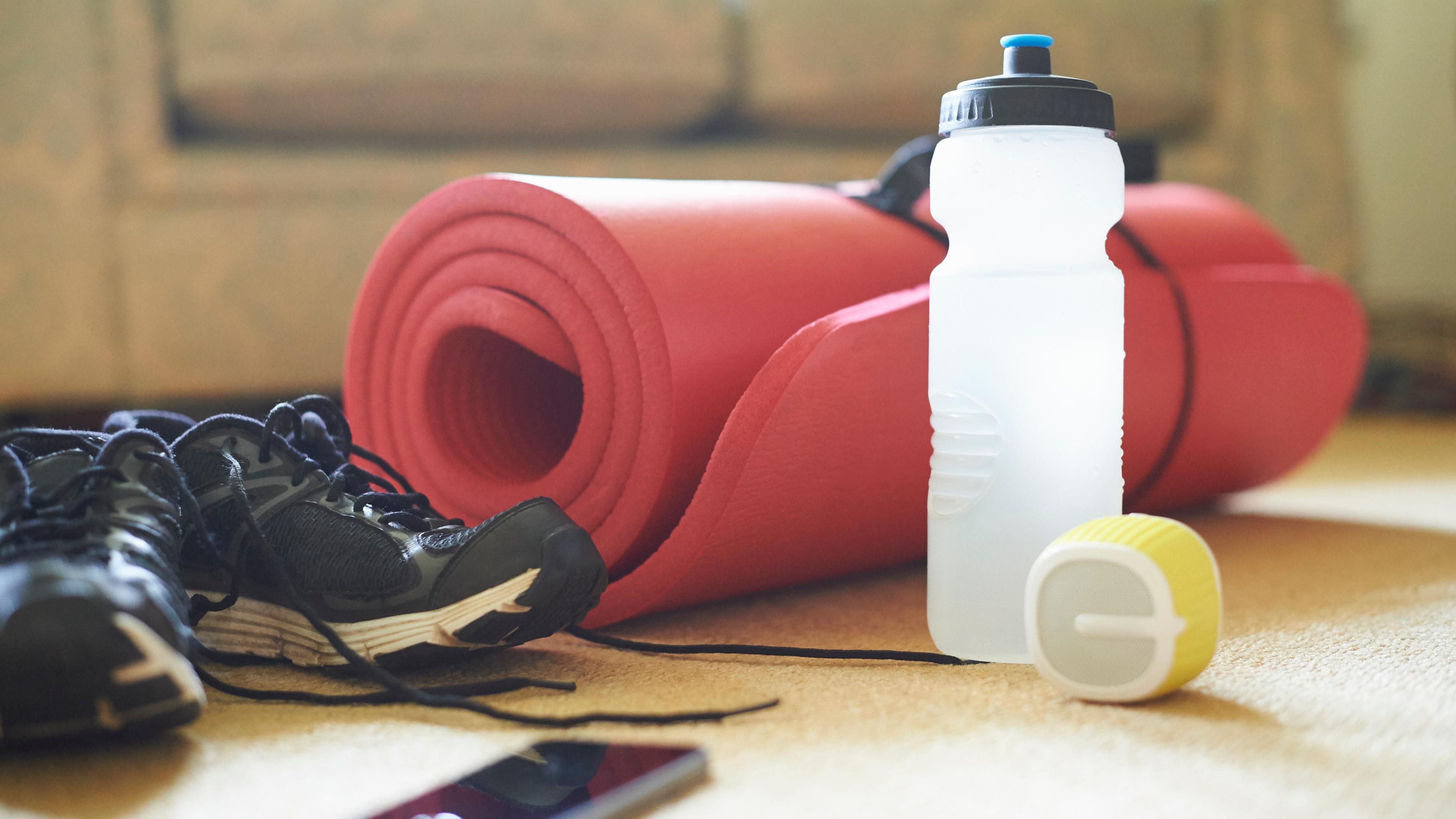 Photo of gym shoes, a red rolled yoga mat, a water bottle and a speaker on a carpeted floor in a cosy room.