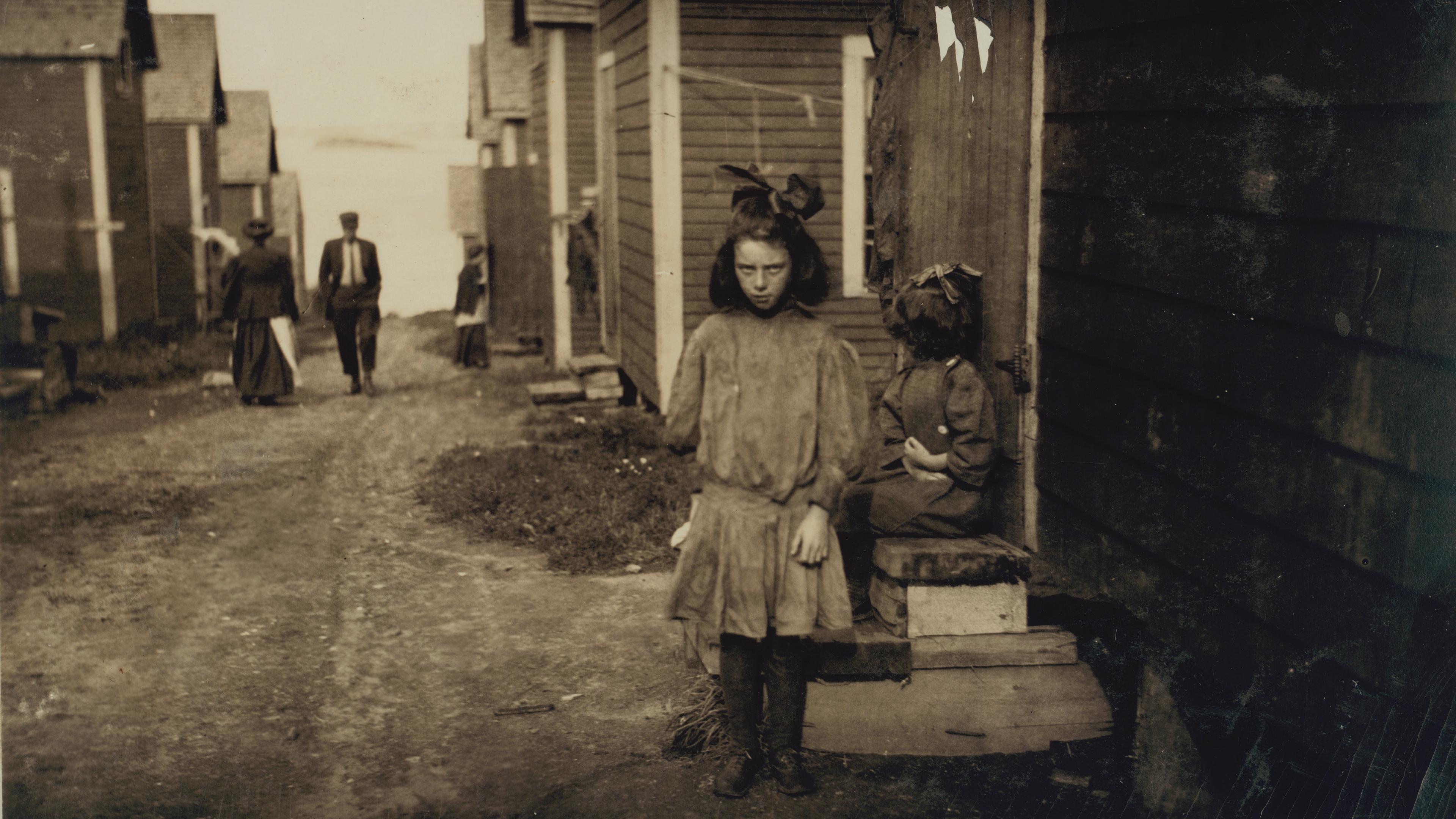 Old photo of a girl standing in a dirt lane between wooden houses with people walking in the background.