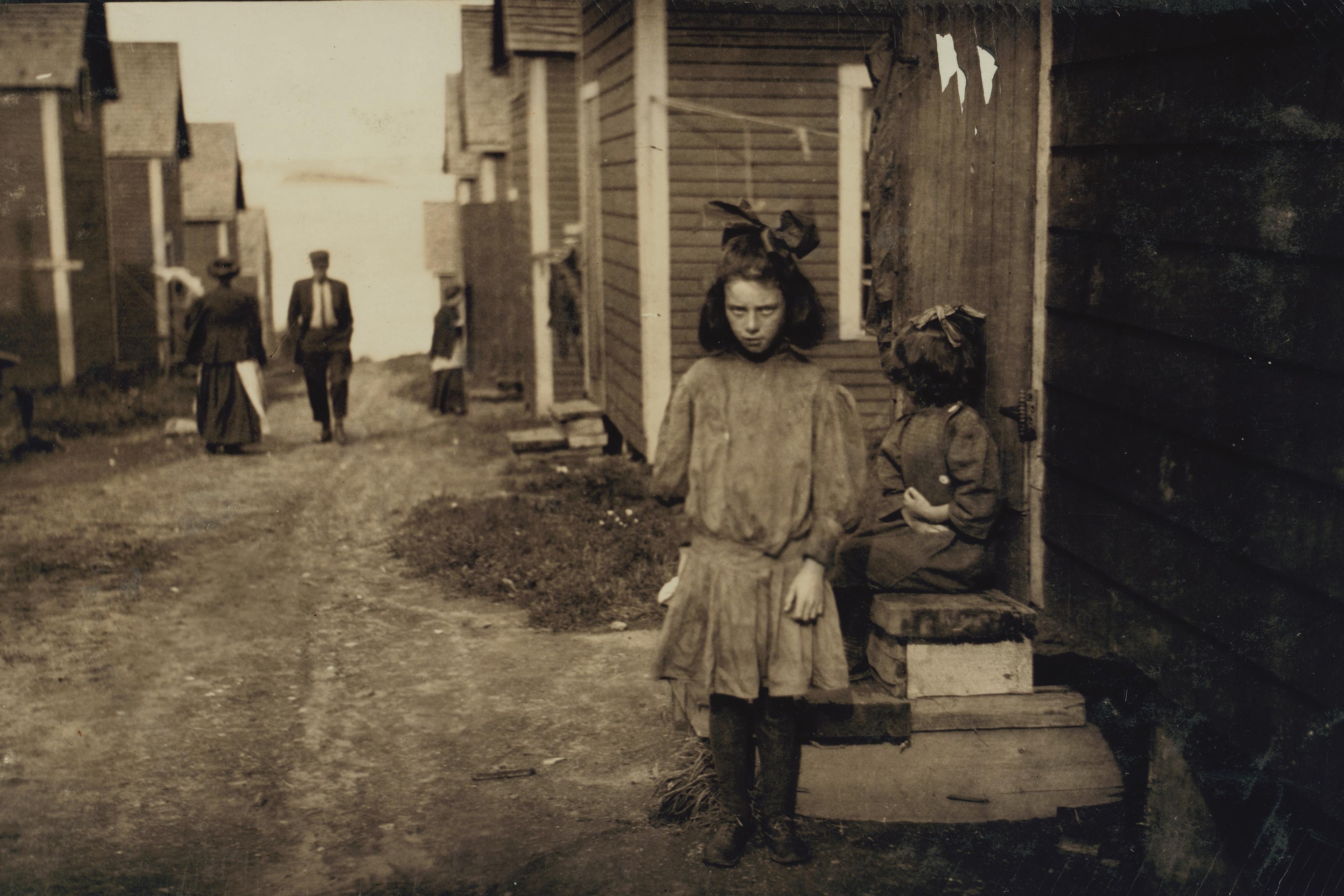 Old photo of a girl standing in a dirt lane between wooden houses with people walking in the background.