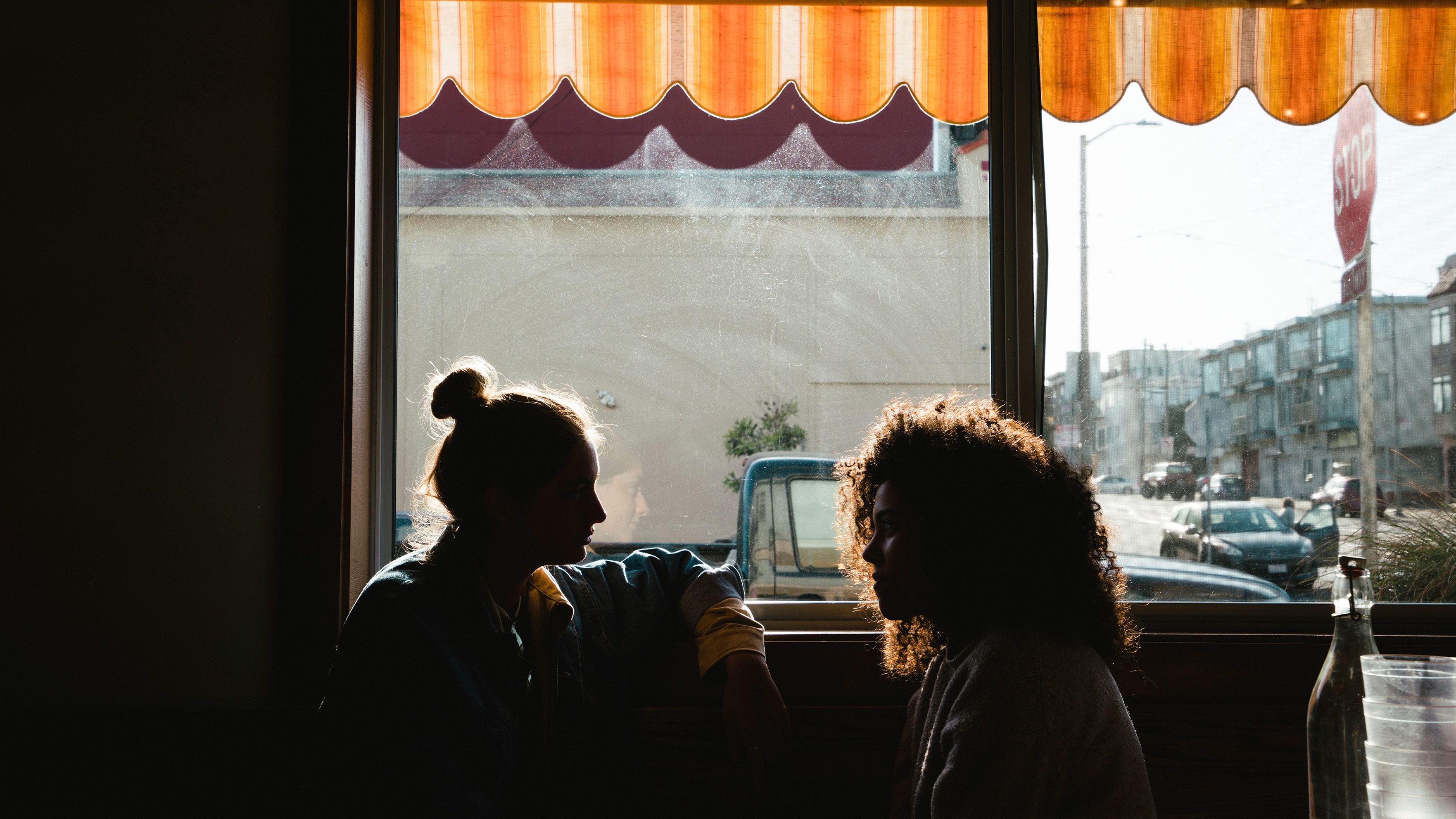 Silhouetted photo of two people talking by a window with sunlight streaming in; buildings and cars visible outside.