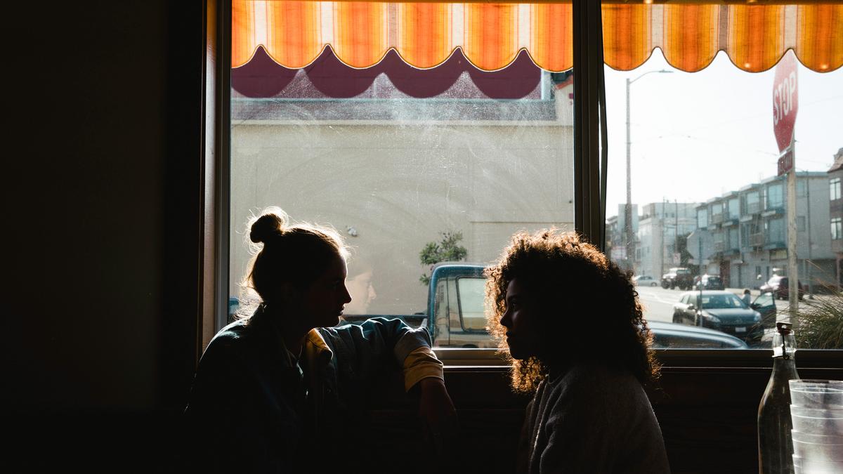 Silhouetted photo of two people talking by a window with sunlight streaming in; buildings and cars visible outside.