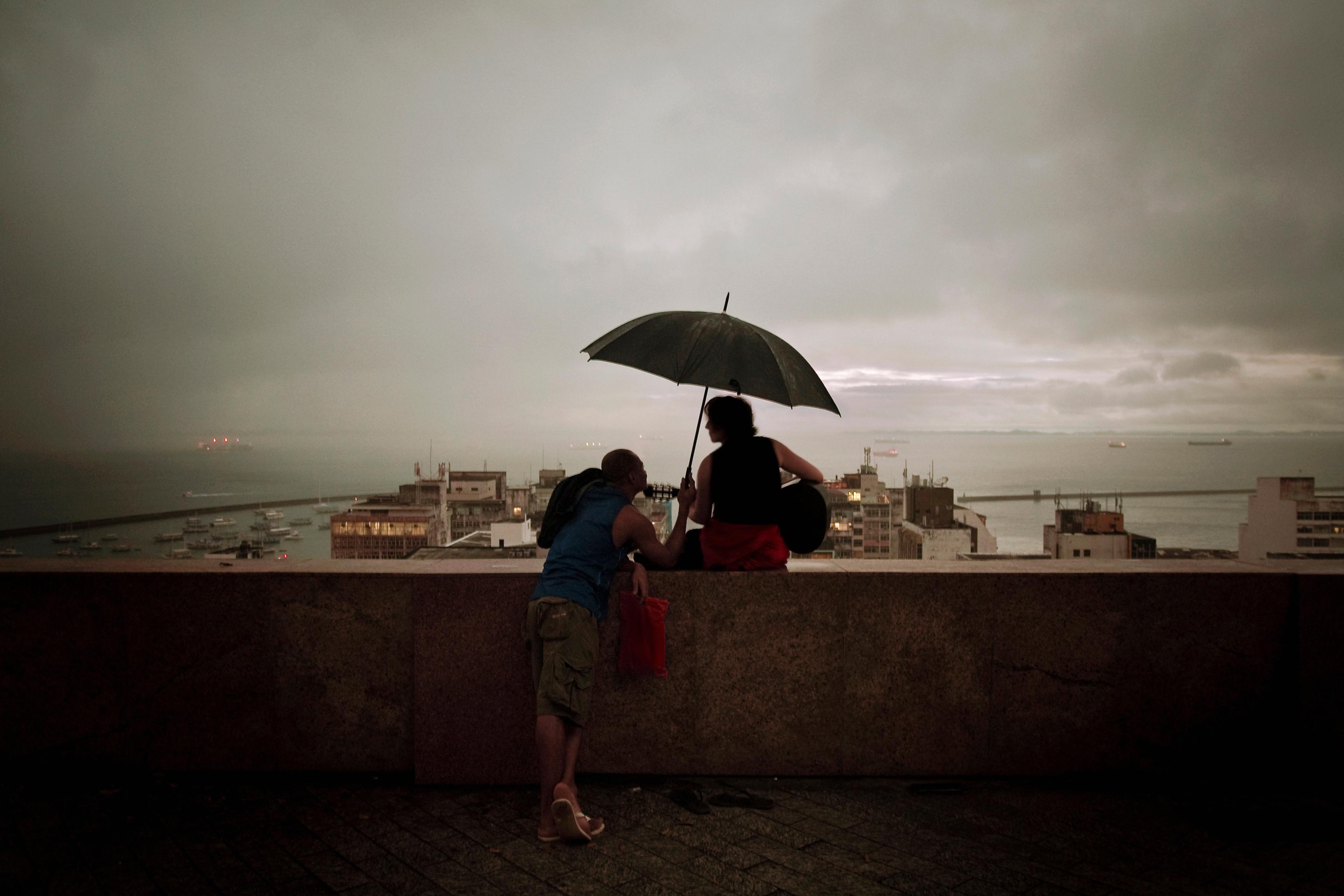 Photo of two people on a ledge under an umbrella during dusk overlooking a cityscape and the sea.