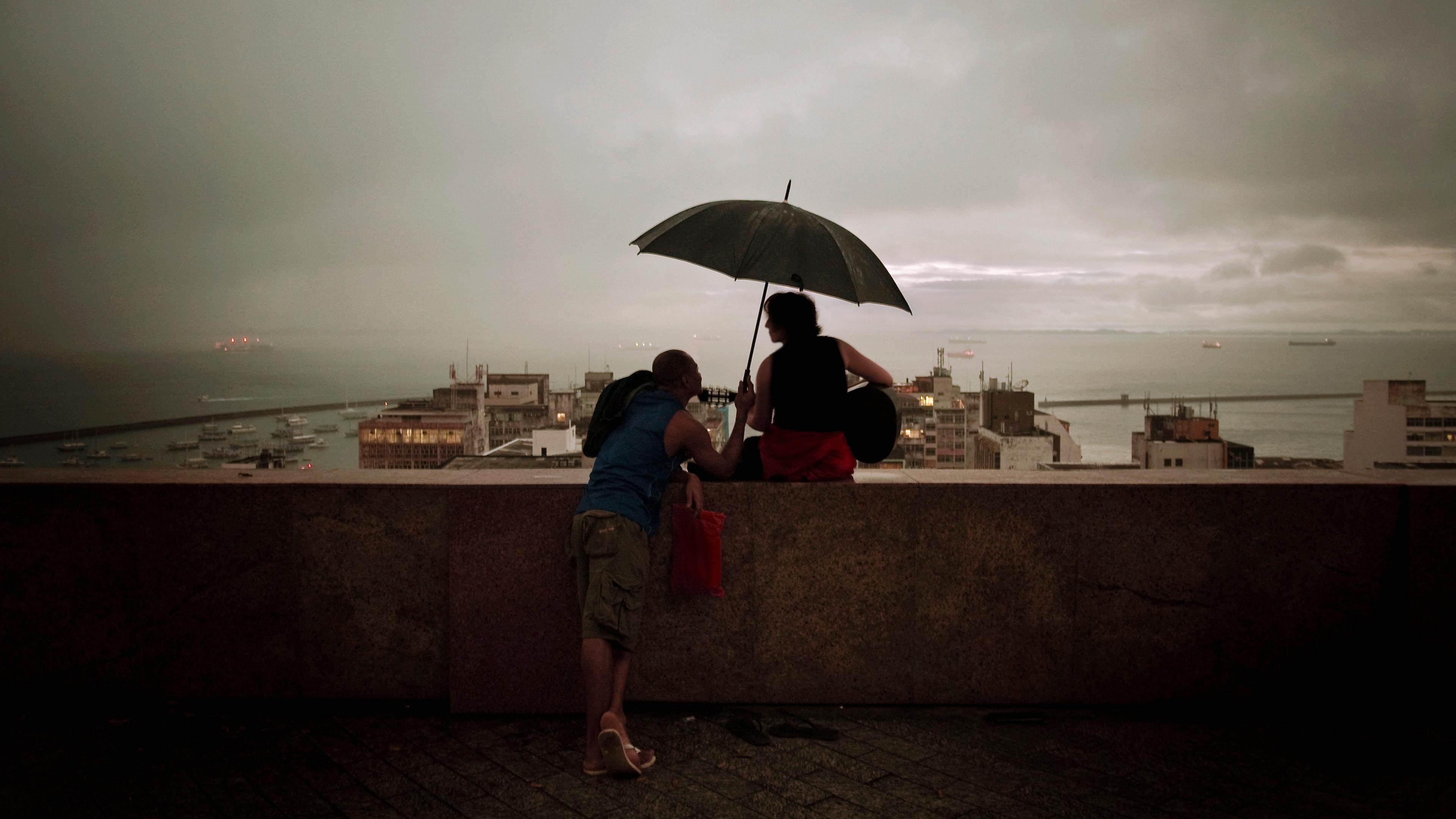 Photo of two people on a ledge under an umbrella during dusk overlooking a cityscape and the sea.