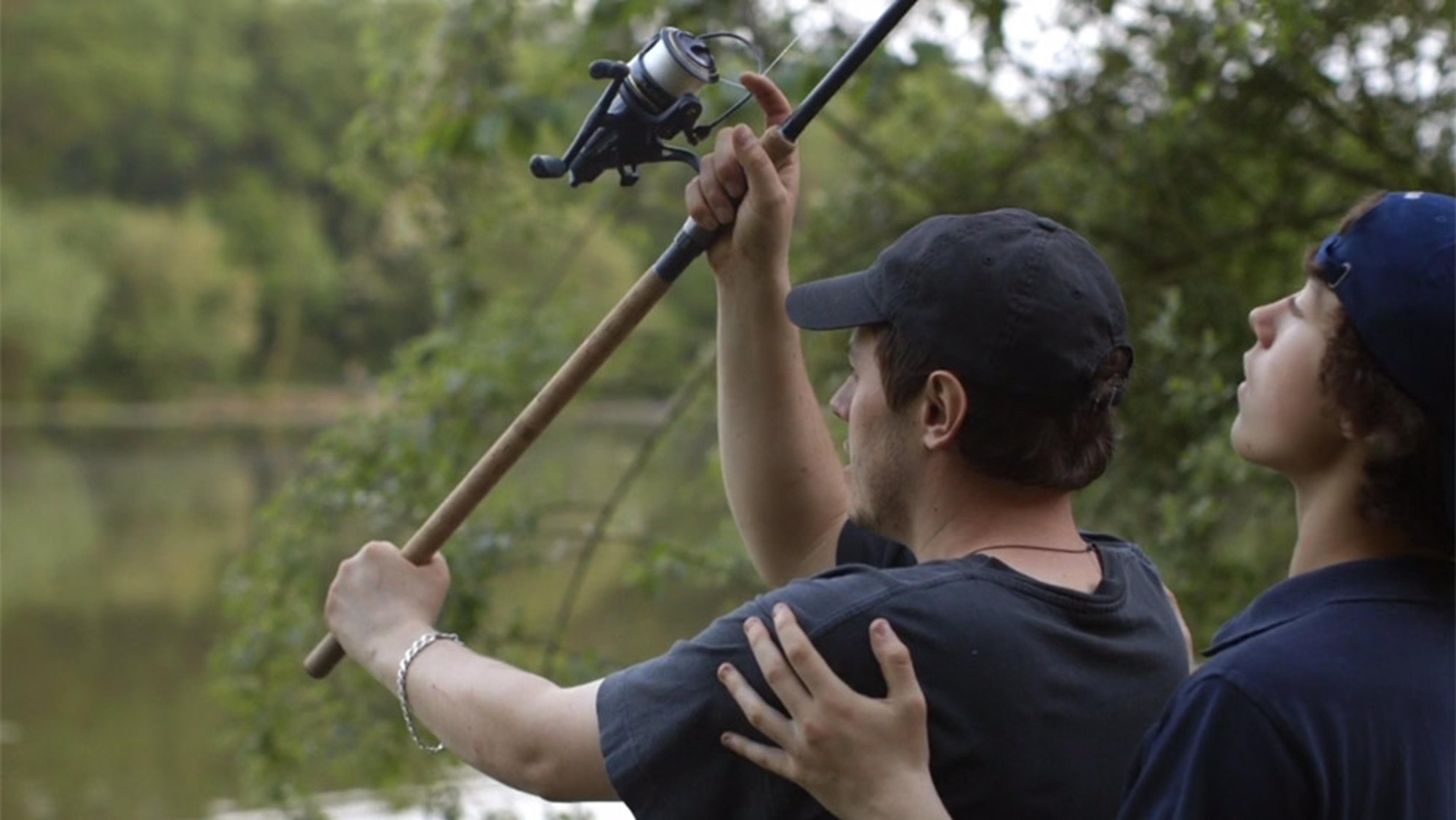 Two people fishing by a lake, one holding a fishing rod while the other assists, surrounded by trees in the background.