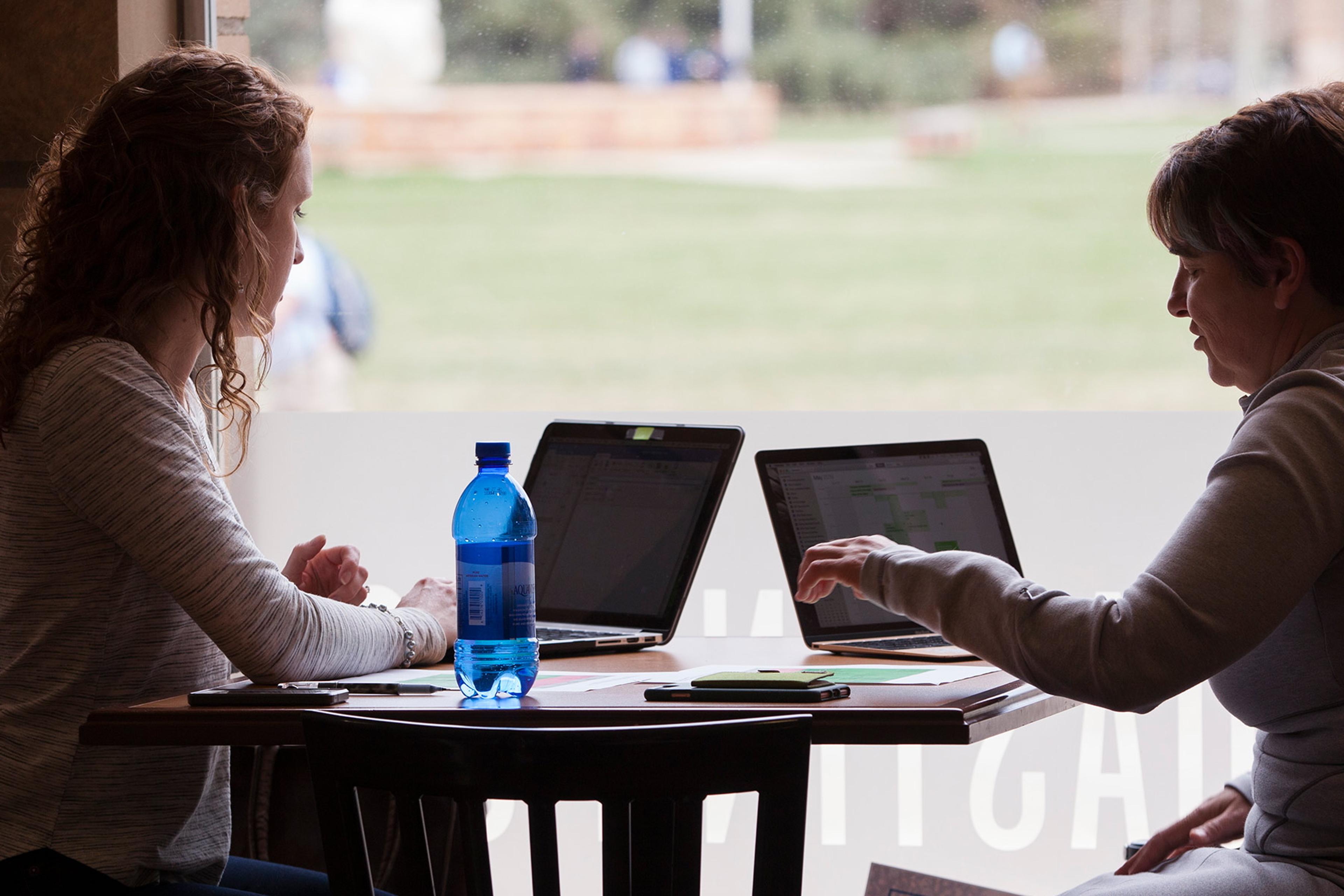 Photo of two people working on laptops at a table indoors, with a bottle of water and blurred outdoor background.