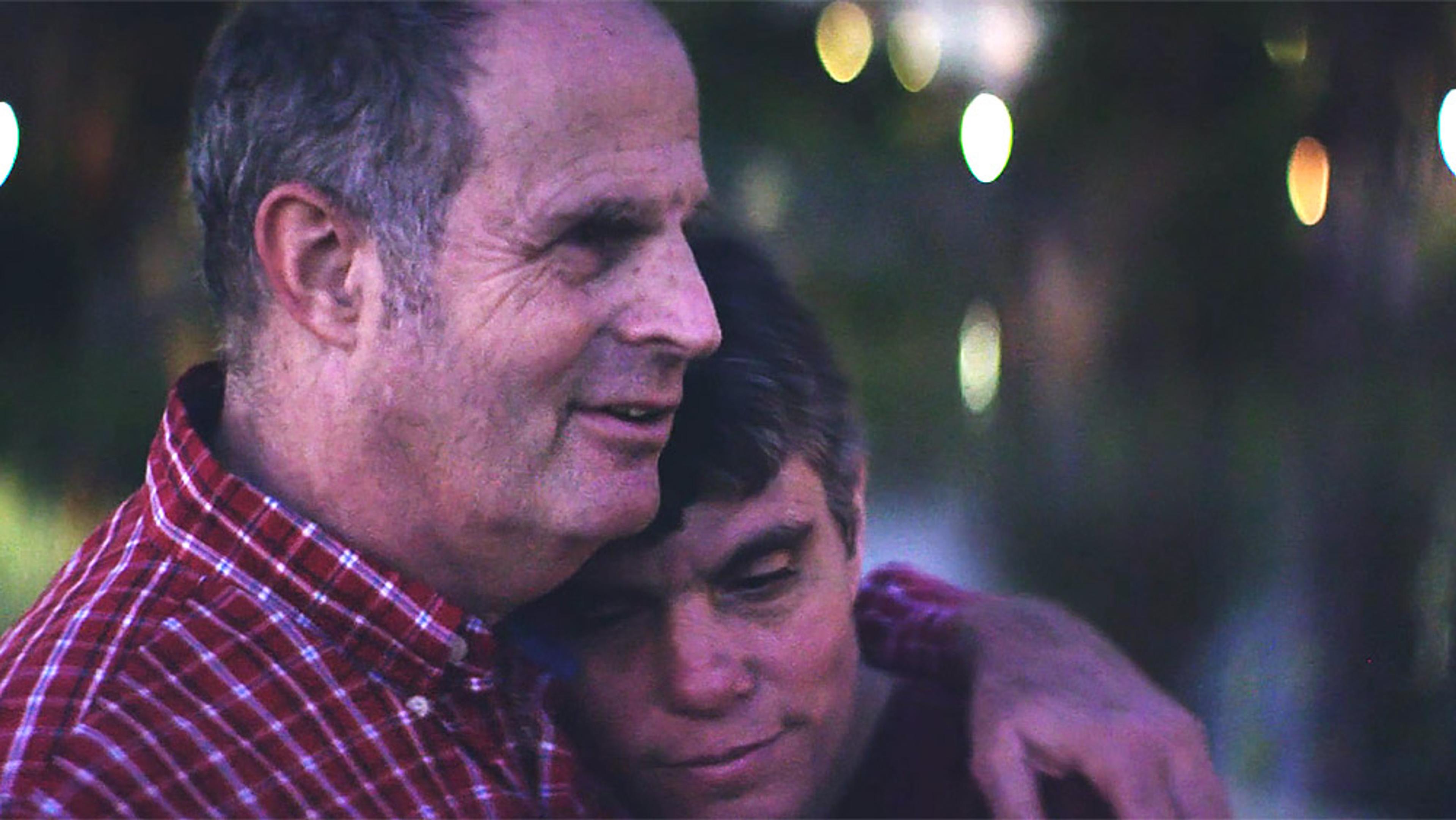A smiling older man in a red plaid shirt hugging a younger person outdoors at night with blurred lights in the background.