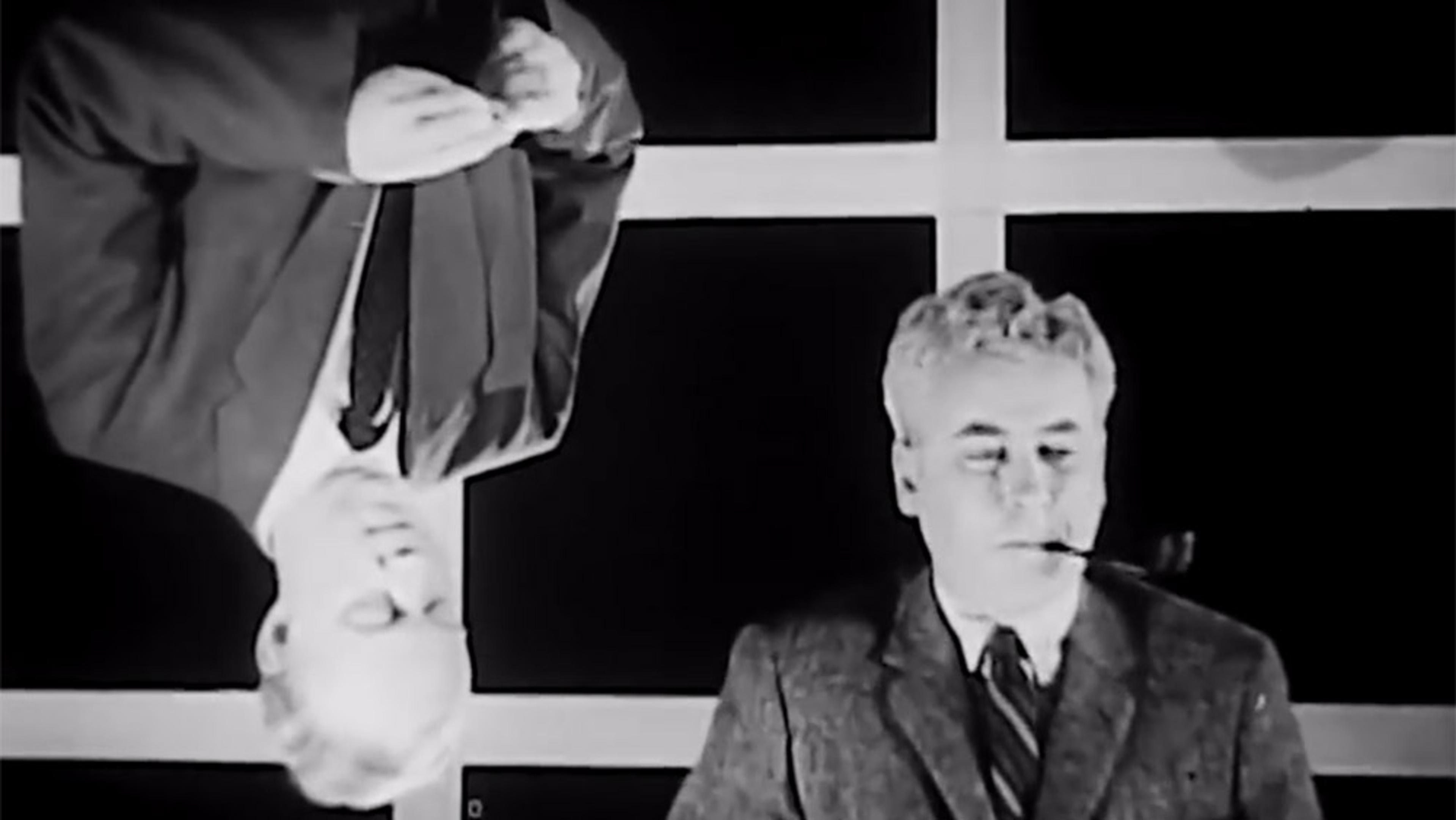 Black-and-white photo of two men in suits against a grid window background, one inverted, the other smoking a pipe.