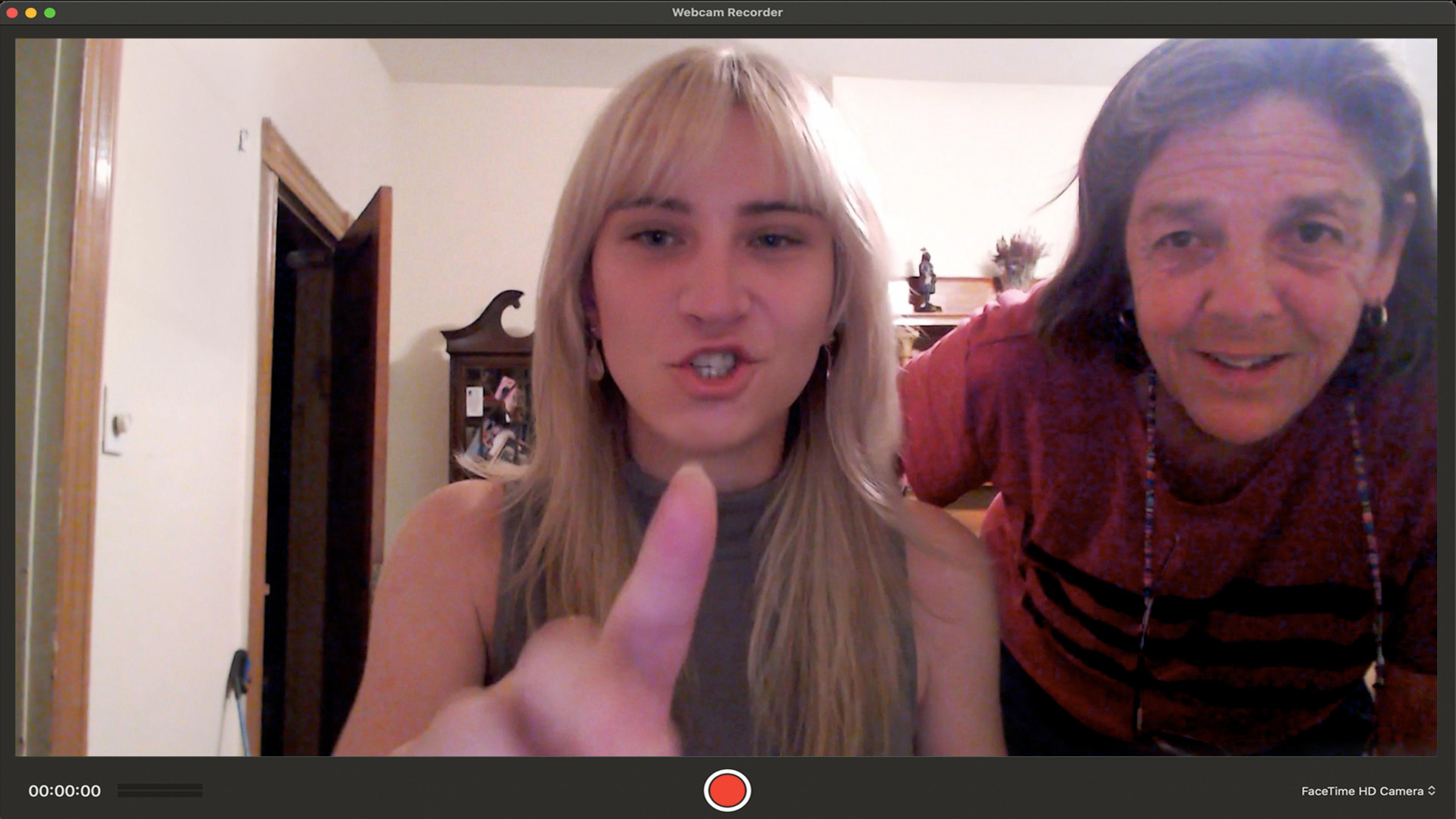 A young woman and an older woman in a living room face the camera, one of them pointing towards it. The image has a webcam recording interface.